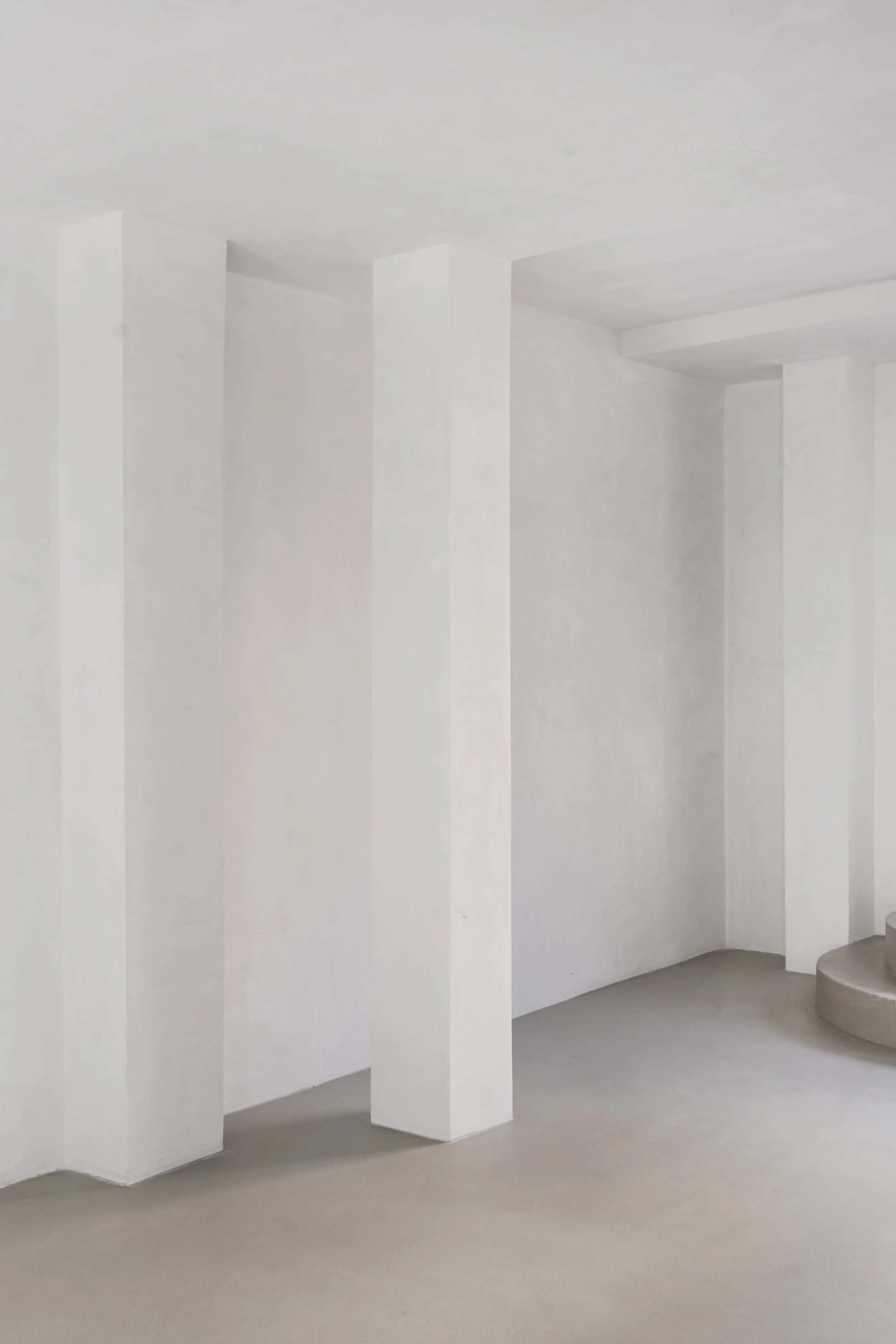 Interior de un espacio arquitectónico con paredes blancas, columnas angulares y una silla circular de concreto en la esquina derecha.