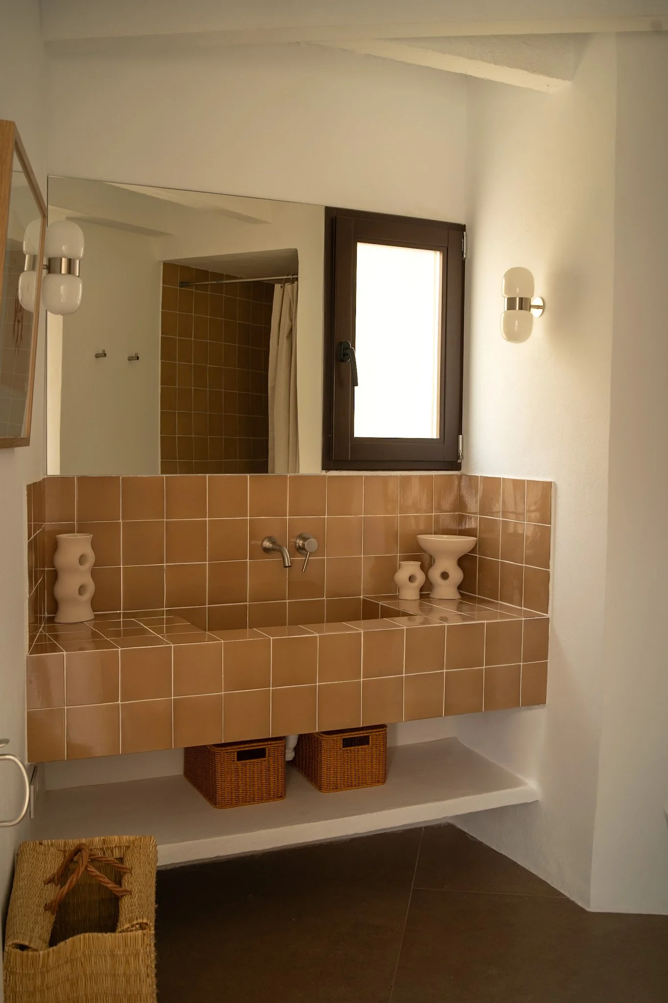 Baño con bañera de azulejos terracota y accesorios decorativos en cerámica, ventana con marco de madera y paredes blancas. Incluye cestas de mimbre y accesorios de baño.
