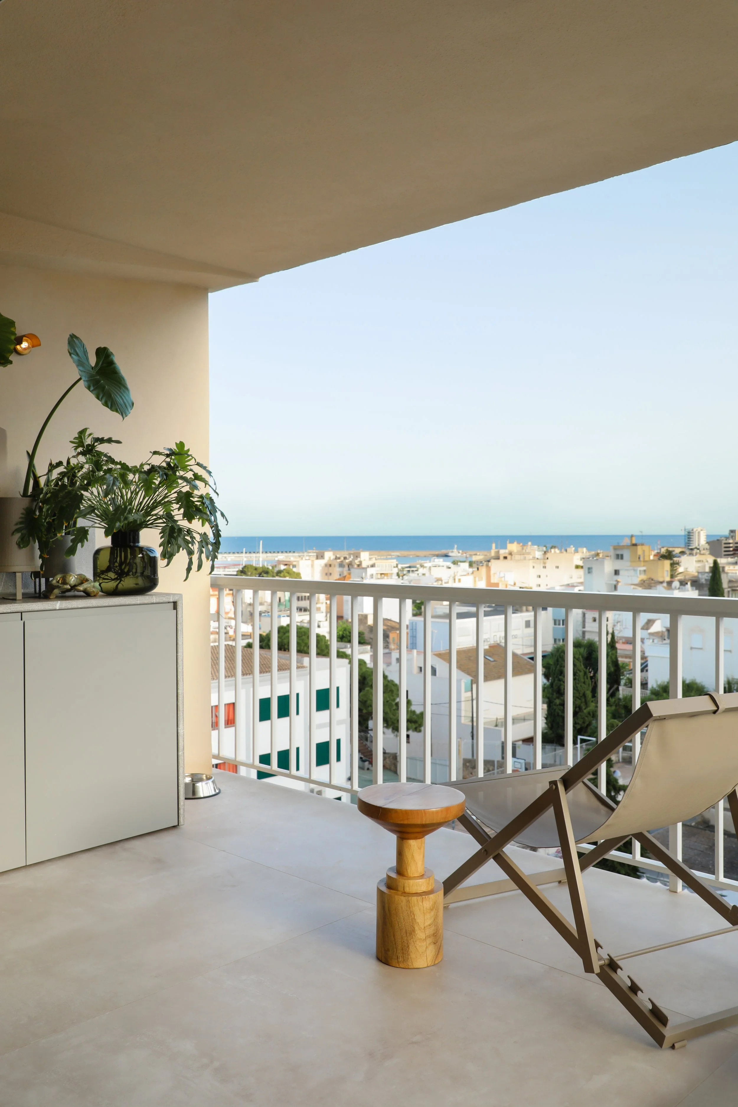 Vista de un balcón con sillas de descanso, una mesa pequeña de madera, plantas en una olla y vista al mar y ciudad bajo un cielo despejado.