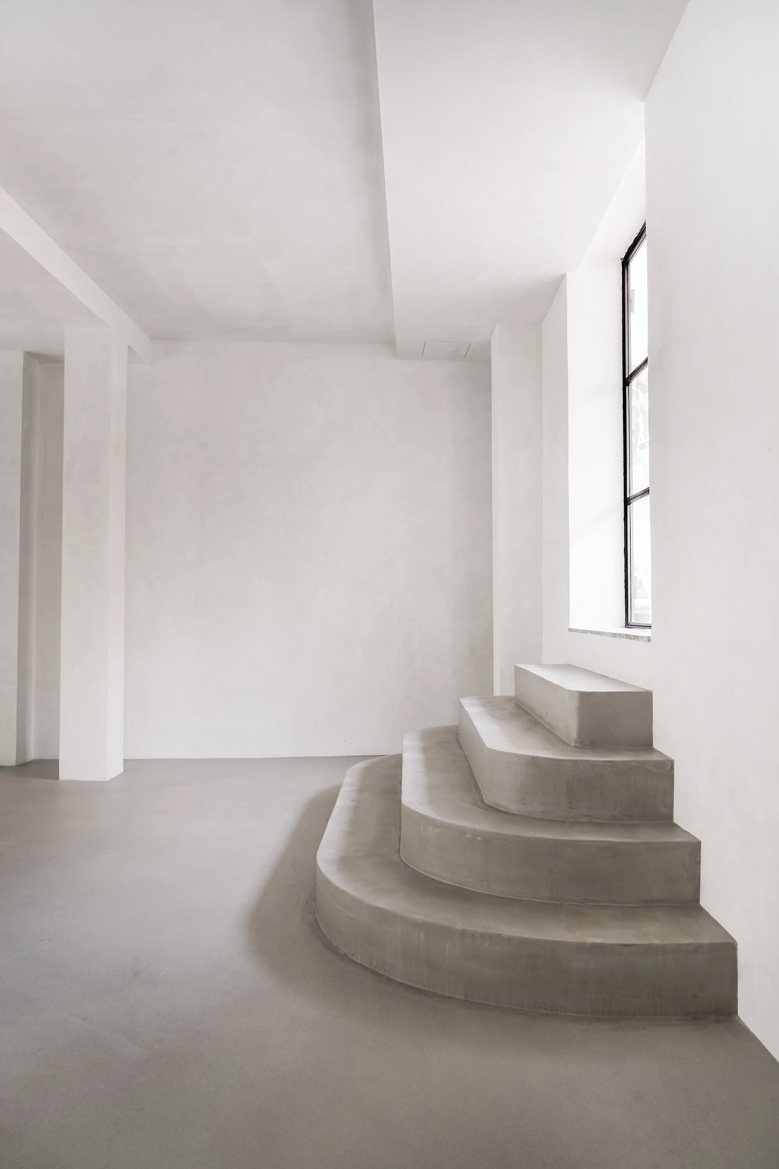 Interior minimalista con escaleras de concreto, paredes blancas y ventanas altas que dejan entrar luz natural.