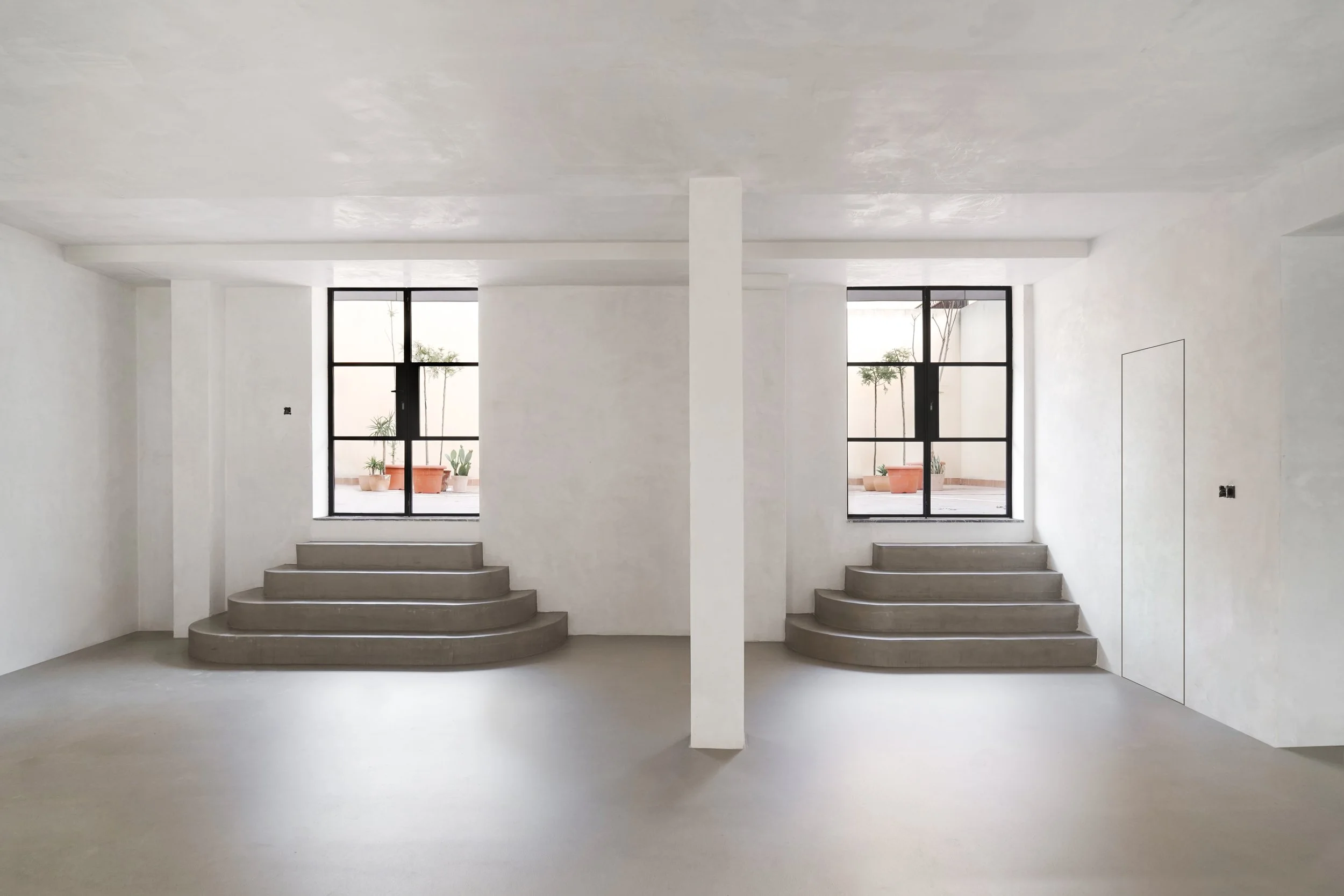 Interior de un espacio minimalista con paredes blancas, ventanas grandes y plantas en la terraza exterior.