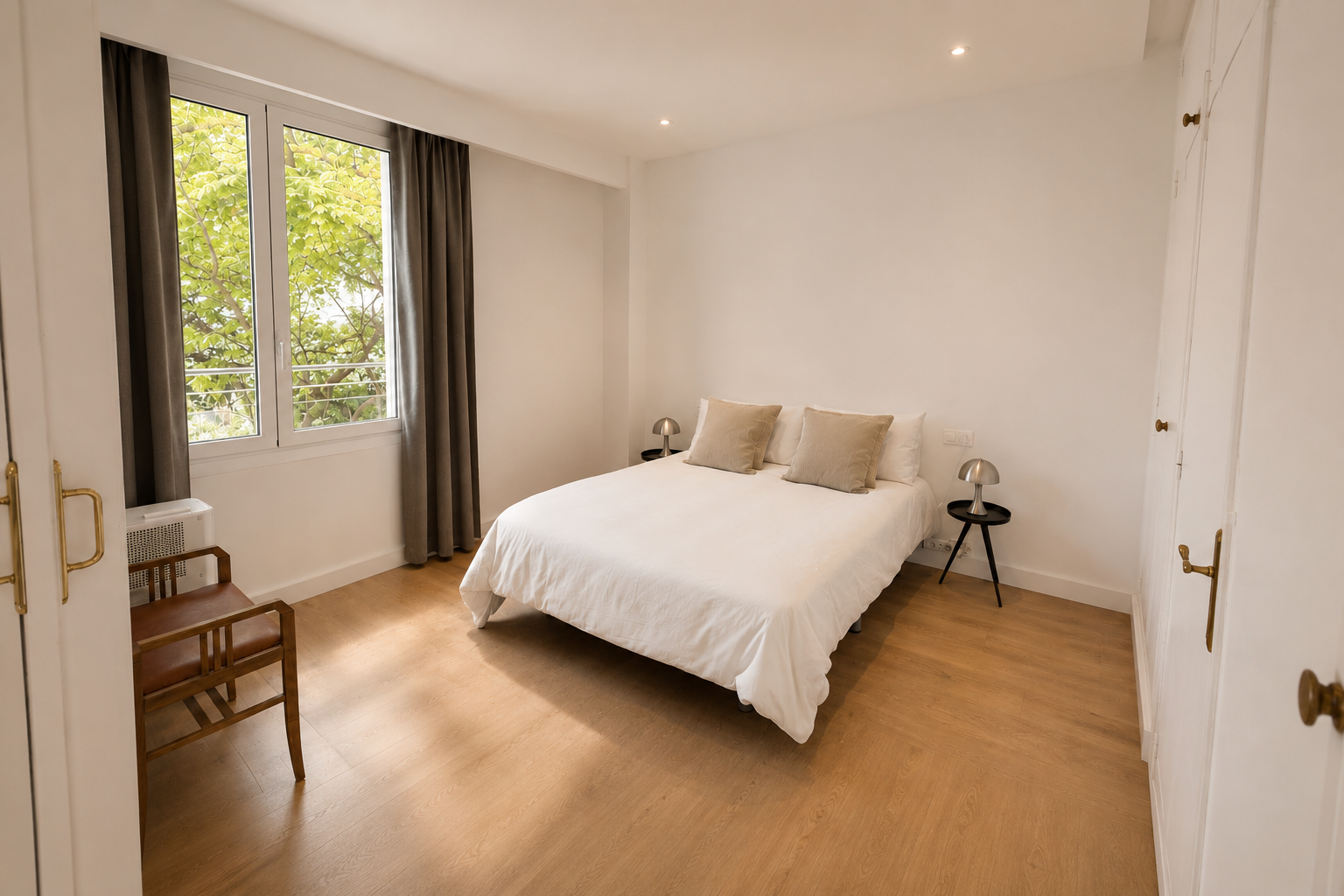 Habitación con cama de matrimonio, dos mesas de noche con lámparas, sillón de madera en la esquina, ventana con cortinas oscuras y vista a árboles
