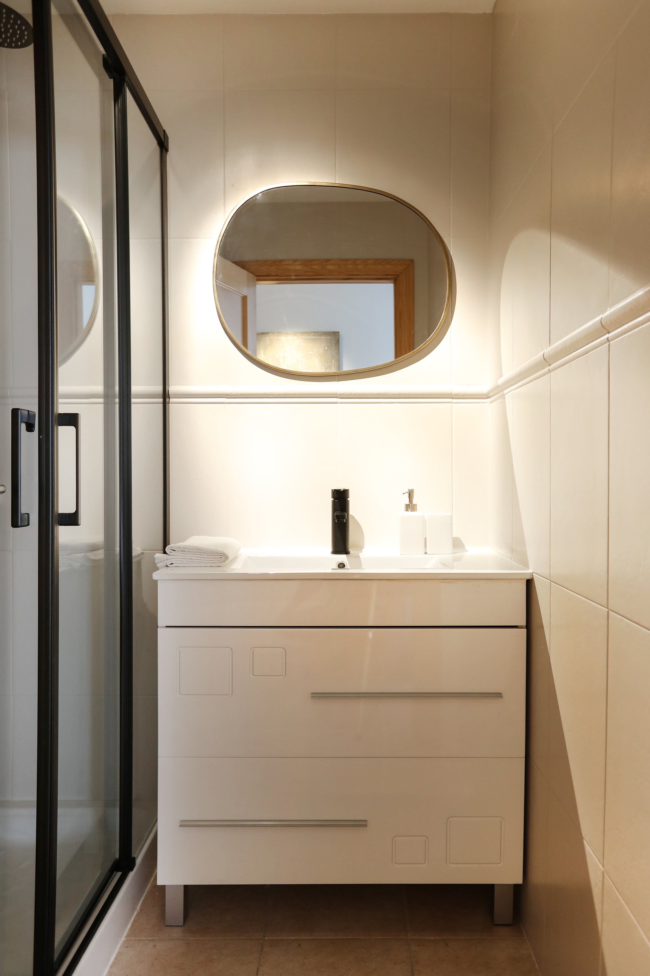Espejo ovalado sobre lavabo blanco con una caja de jabón y toallas dobladas, en un baño de paredes beige y puerta de madera visible en el reflejo.