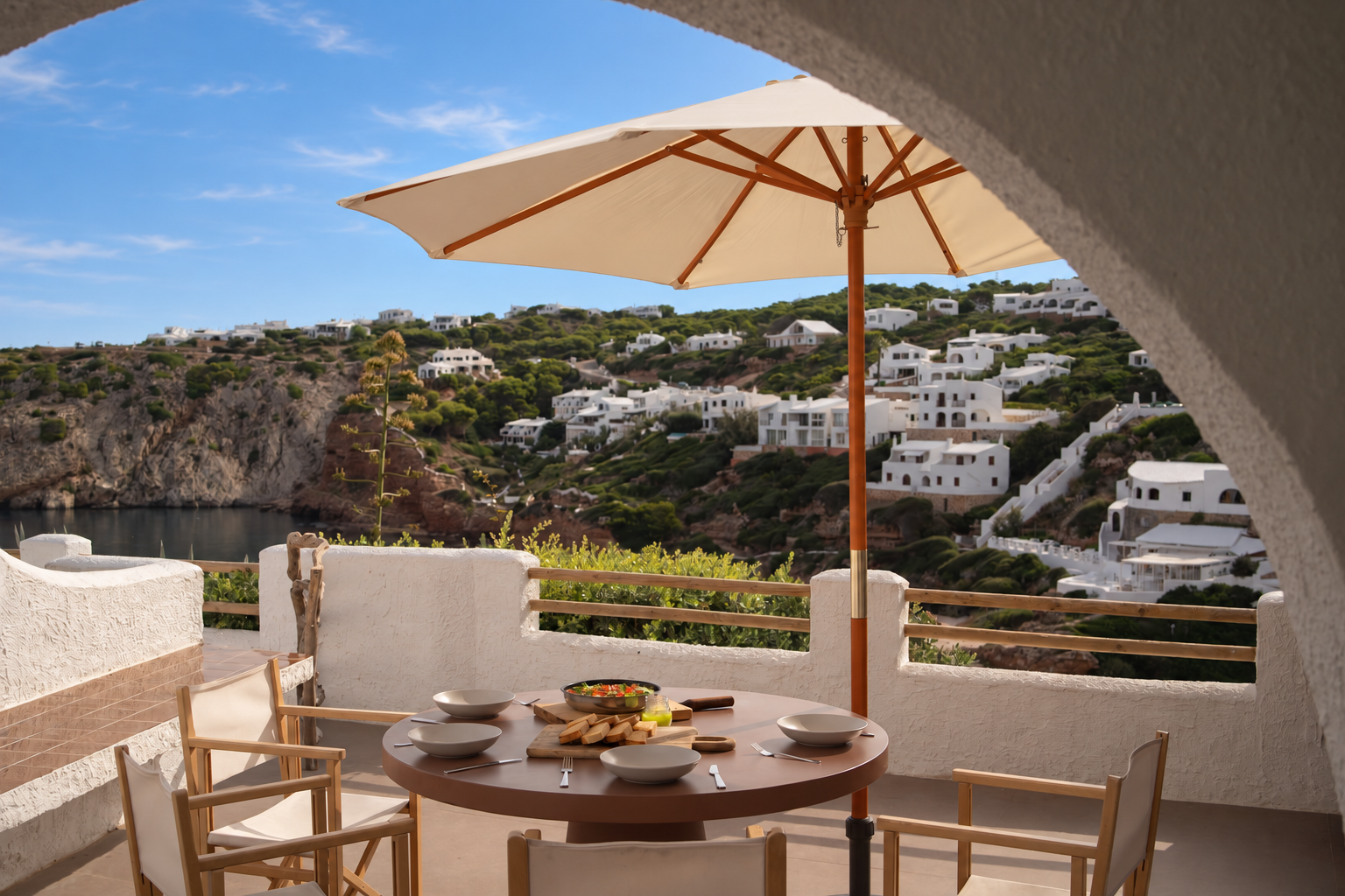 Outdoor patio with a table set for a meal, surrounded by white stucco walls and a large beige umbrella, overlooking a scenic hillside with white buildings and lush greenery.