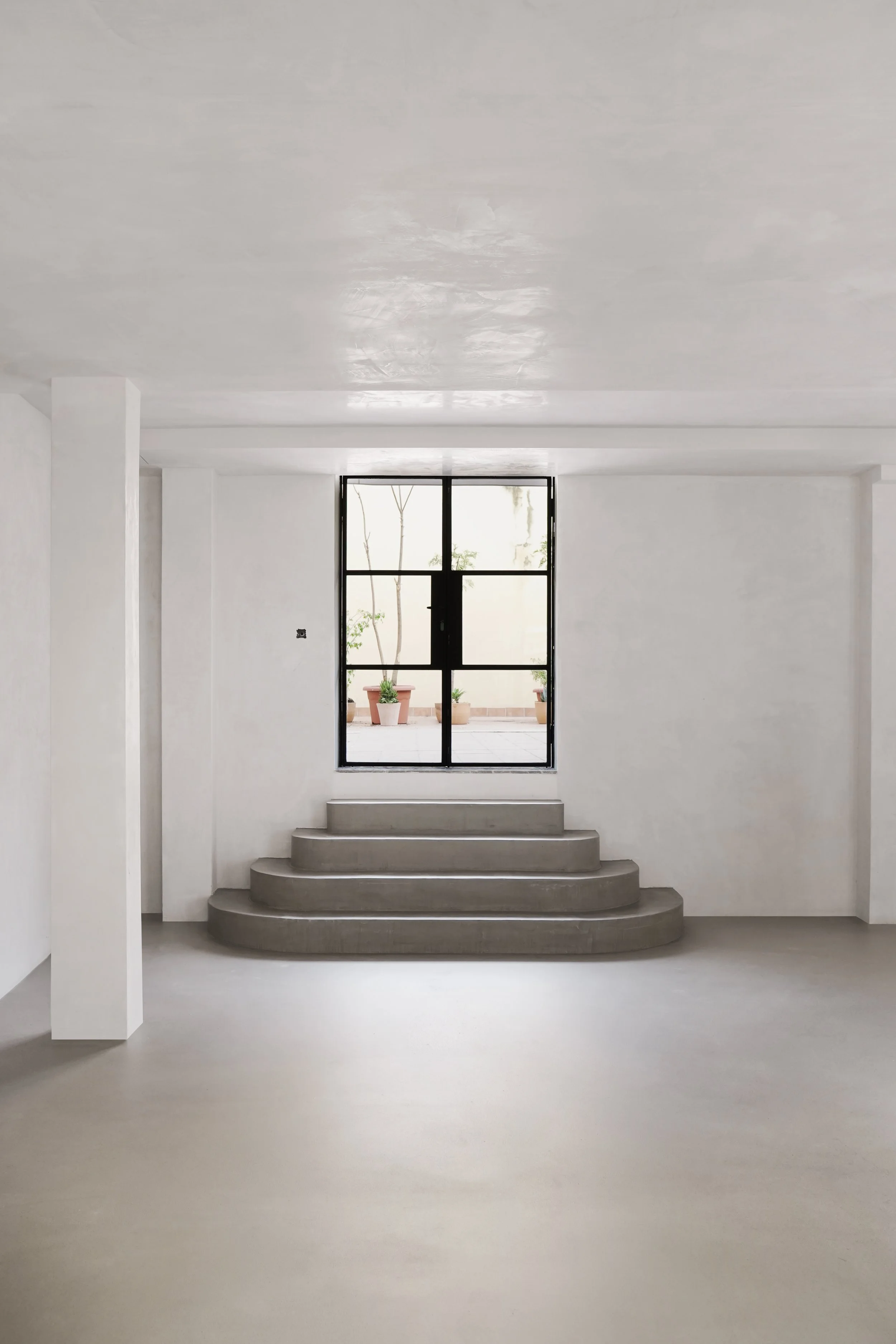 interior de un espacio minimalista con escaleras de concreto hacia un área con una ventana grande y plantas en macetas al exterior