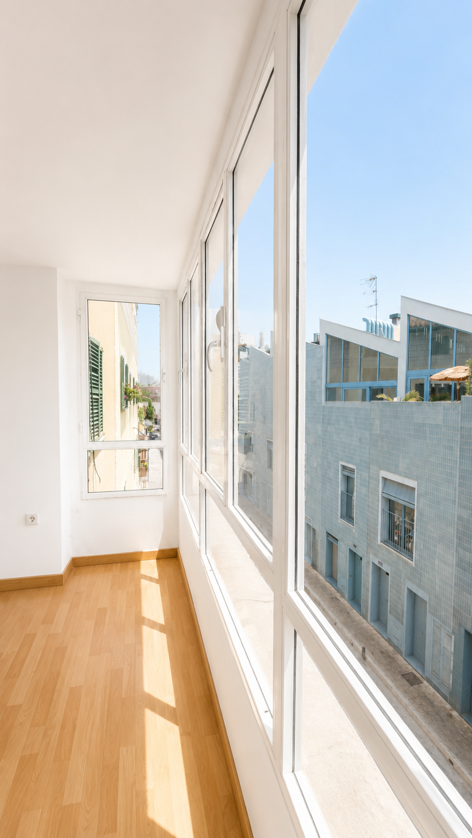 Vista de un balcón cerrado con paredes de ventanas de cristal, suelo de madera clara y edificios urbanos alrededor bajo un cielo despejado.