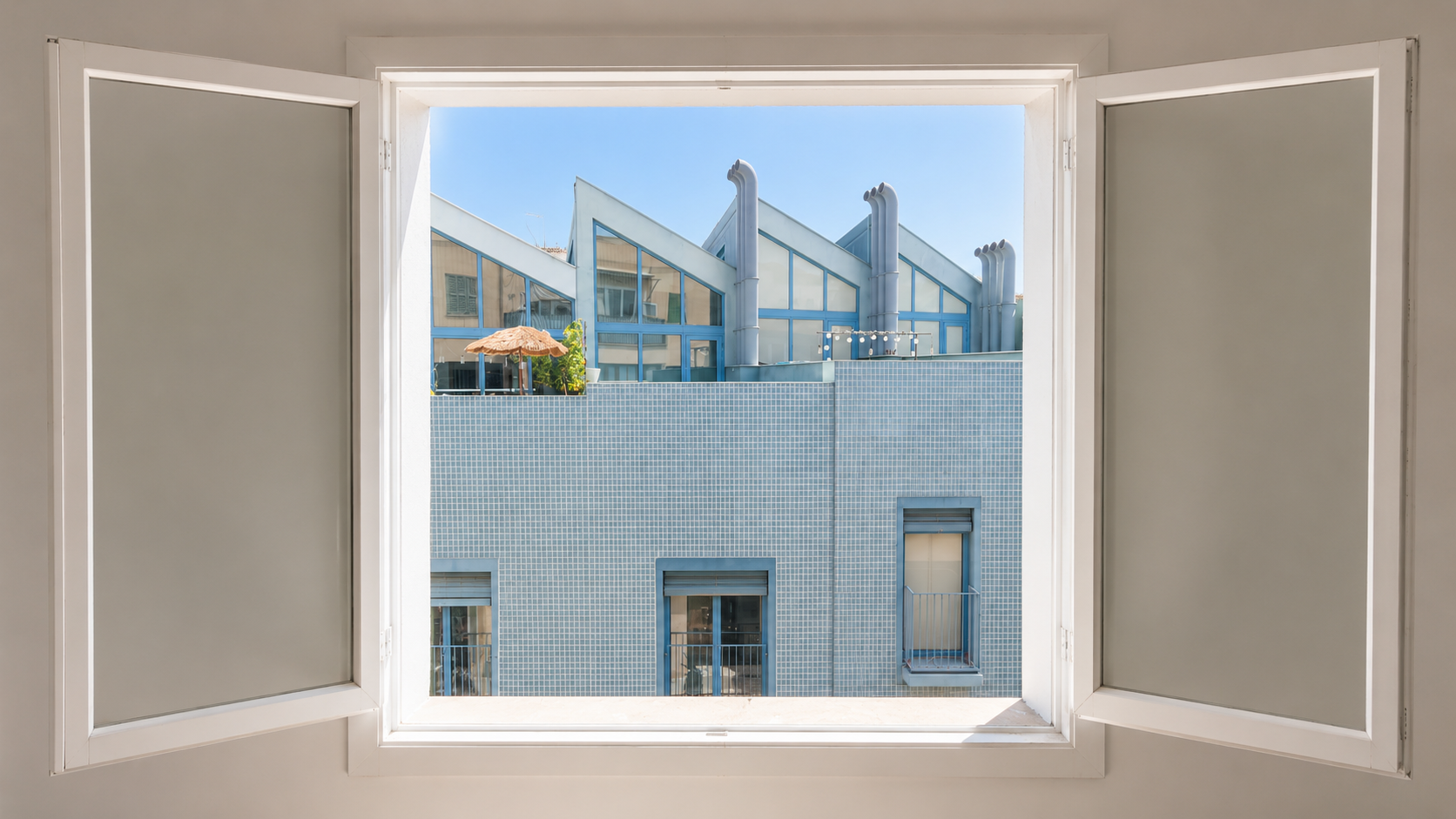 Vista desde una ventana abierta hacia un edificio con fachadas de azulejos azules y ventanas con ventanas corredizas y balcones pequeños. En la azotea hay ventiladores grandes y una sombrilla de paja, con un cielo azul claro al fondo.