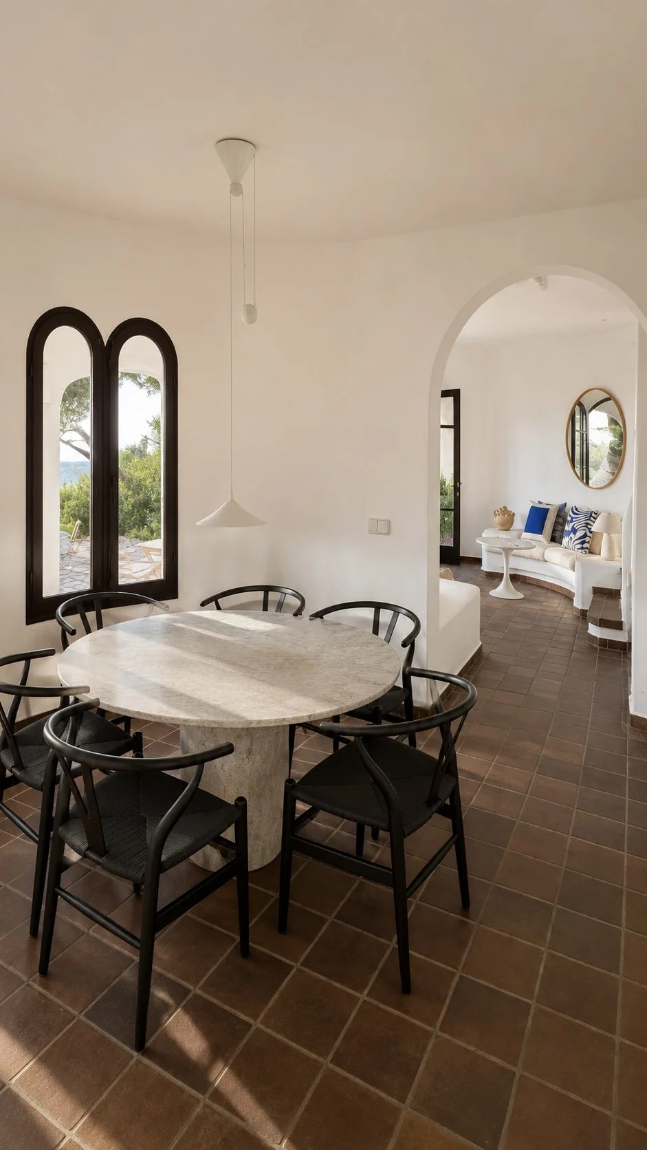Interior de una sala de comedor con mesa de piedra y sillas negras, ventanas de arcos oscuros y vista hacia una terraza exterior con árboles.