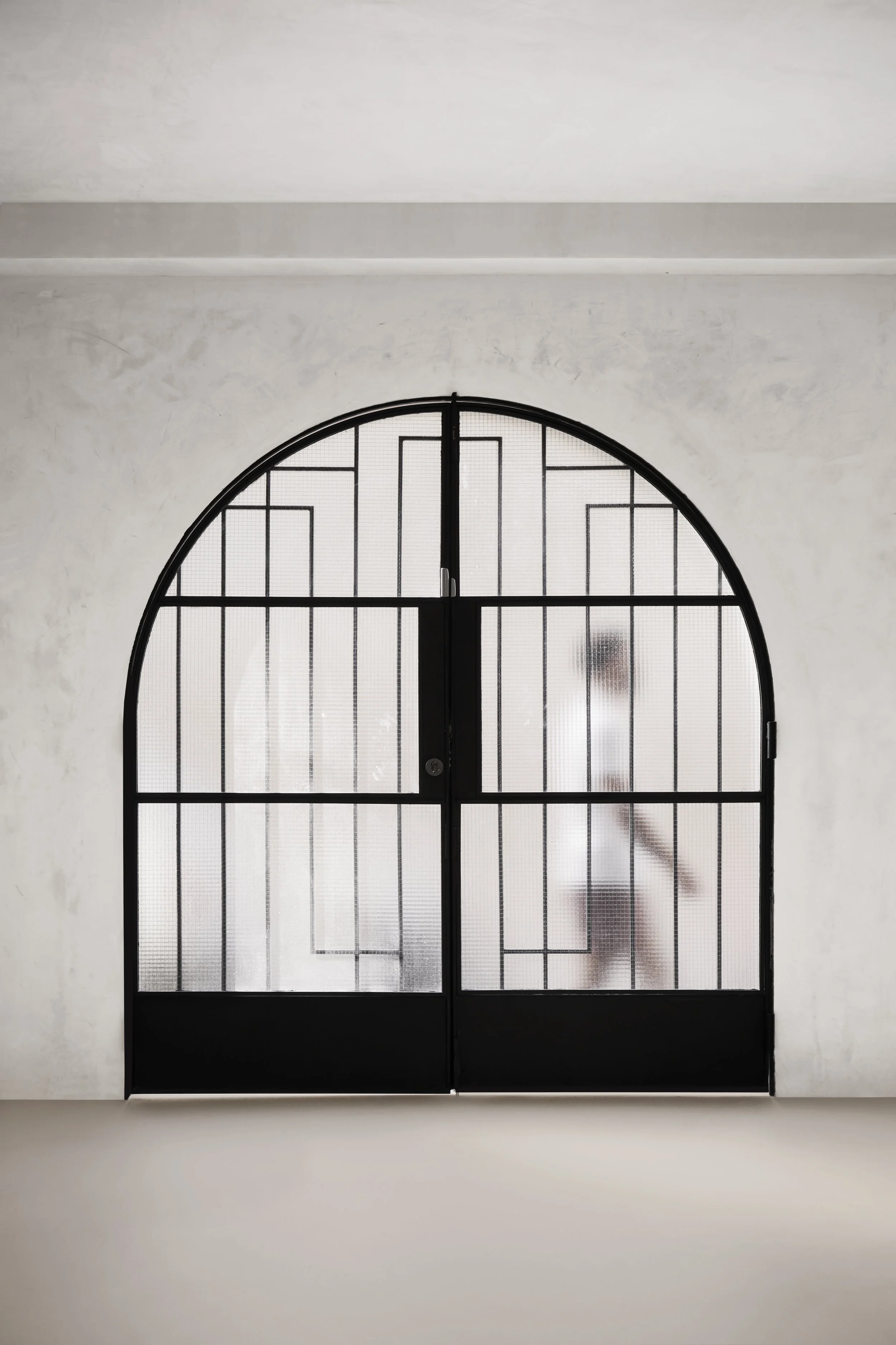 Puerta de metal negra con ventanas de acrílico y marco en forma de arco, cerrada, en una pared blanca.