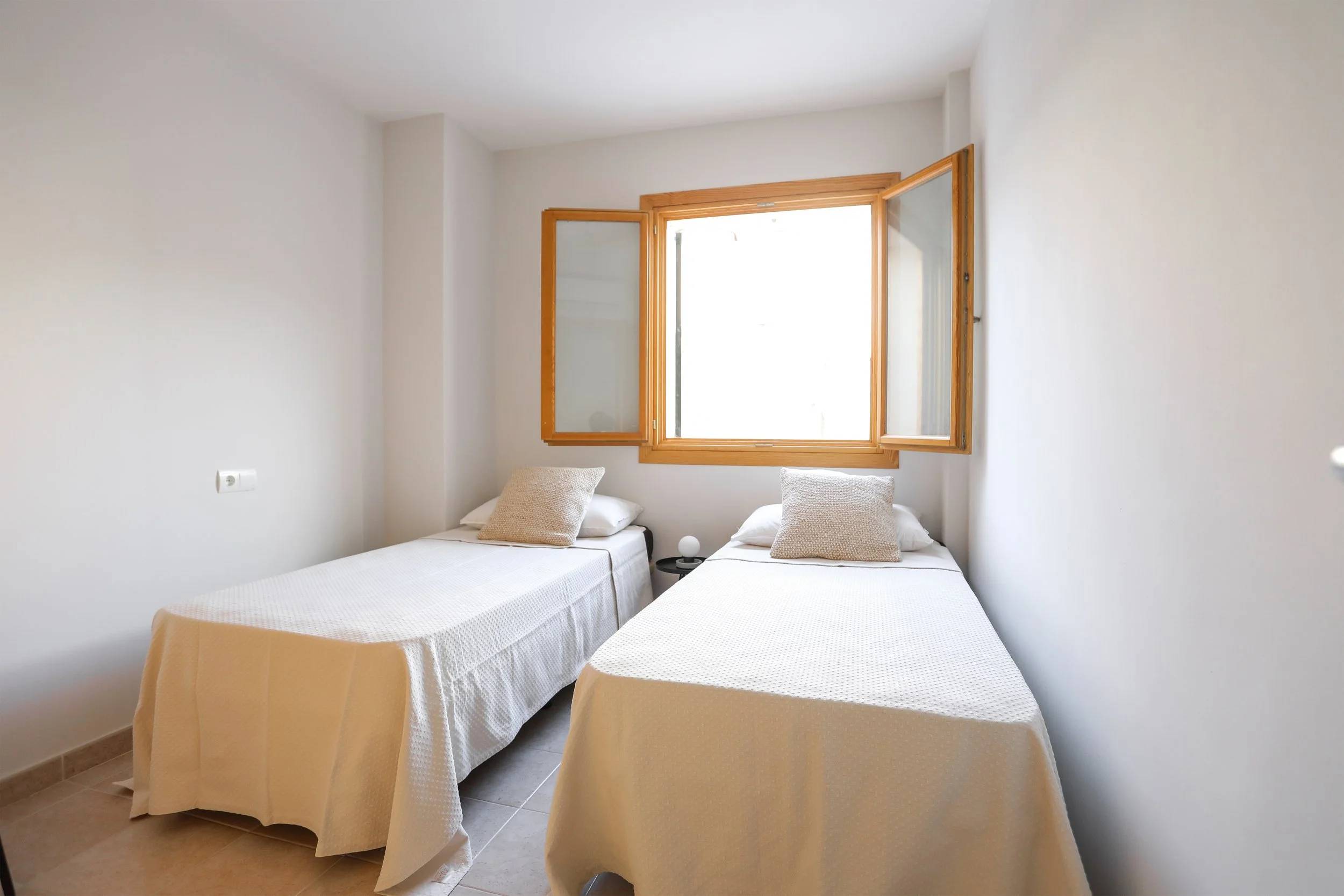 Habitación con dos camas individuales con cubrecasas beige, una ventana de madera con vistas al exterior, paredes blancas y un enchufe eléctrico en la pared izquierda.