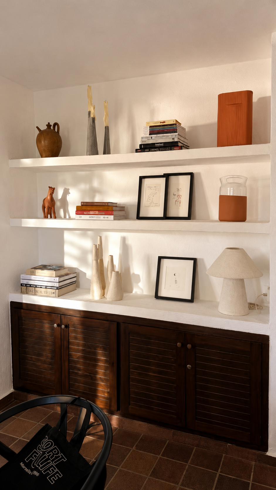 Estantería blanca con objetos decorativos, incluyendo libros, cuadros, cerámicas y una lámpara de cerámica en una sala con suelo de baldosas.