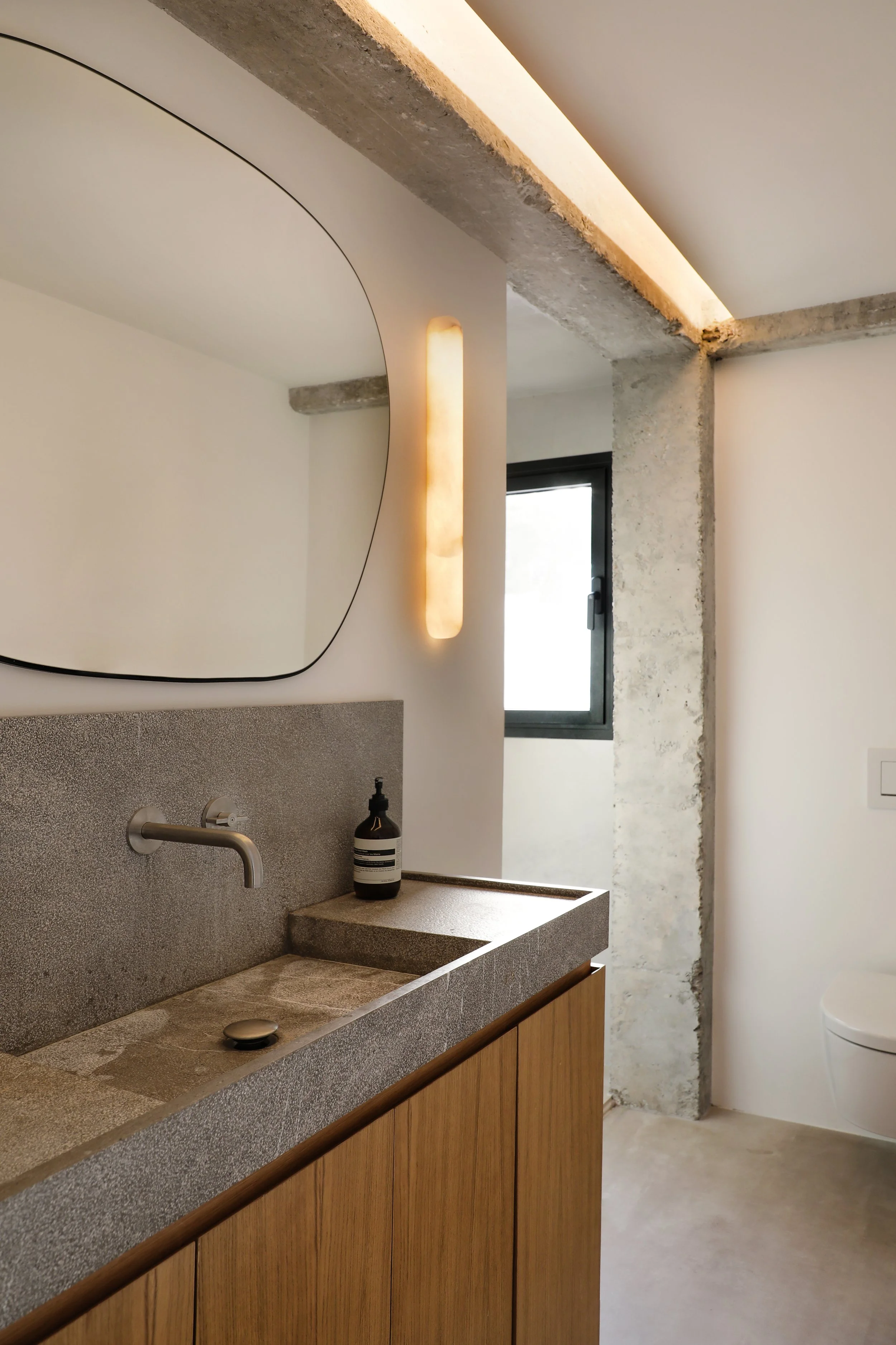 Lavabo de piedra con grifo de metal, espejo redondo y pared de cemento sin acabar en un baño moderno.