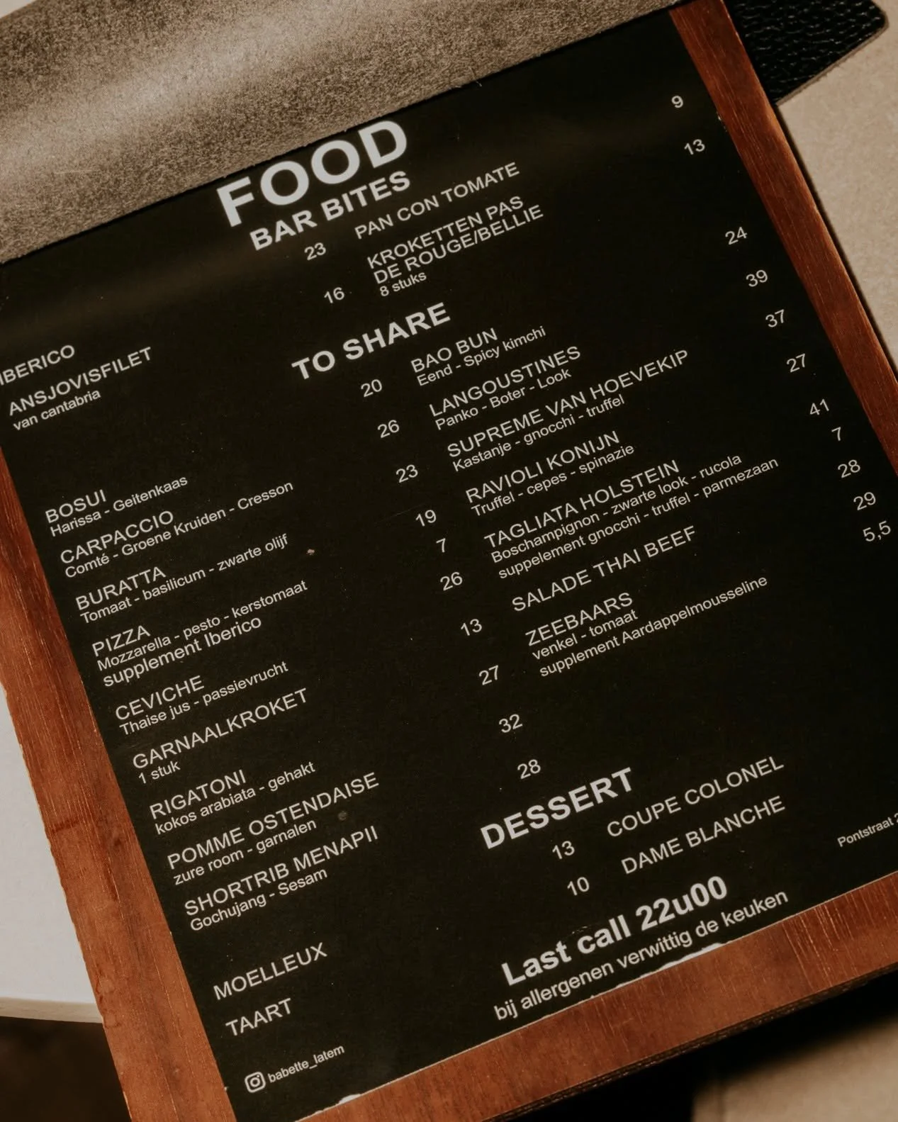 De menukaart van een restaurant met gerechten zoals bar bites, sharing planken, salades, desserts en schotels, geschreven in het Nederlands op een zwart bord met een houten rand.