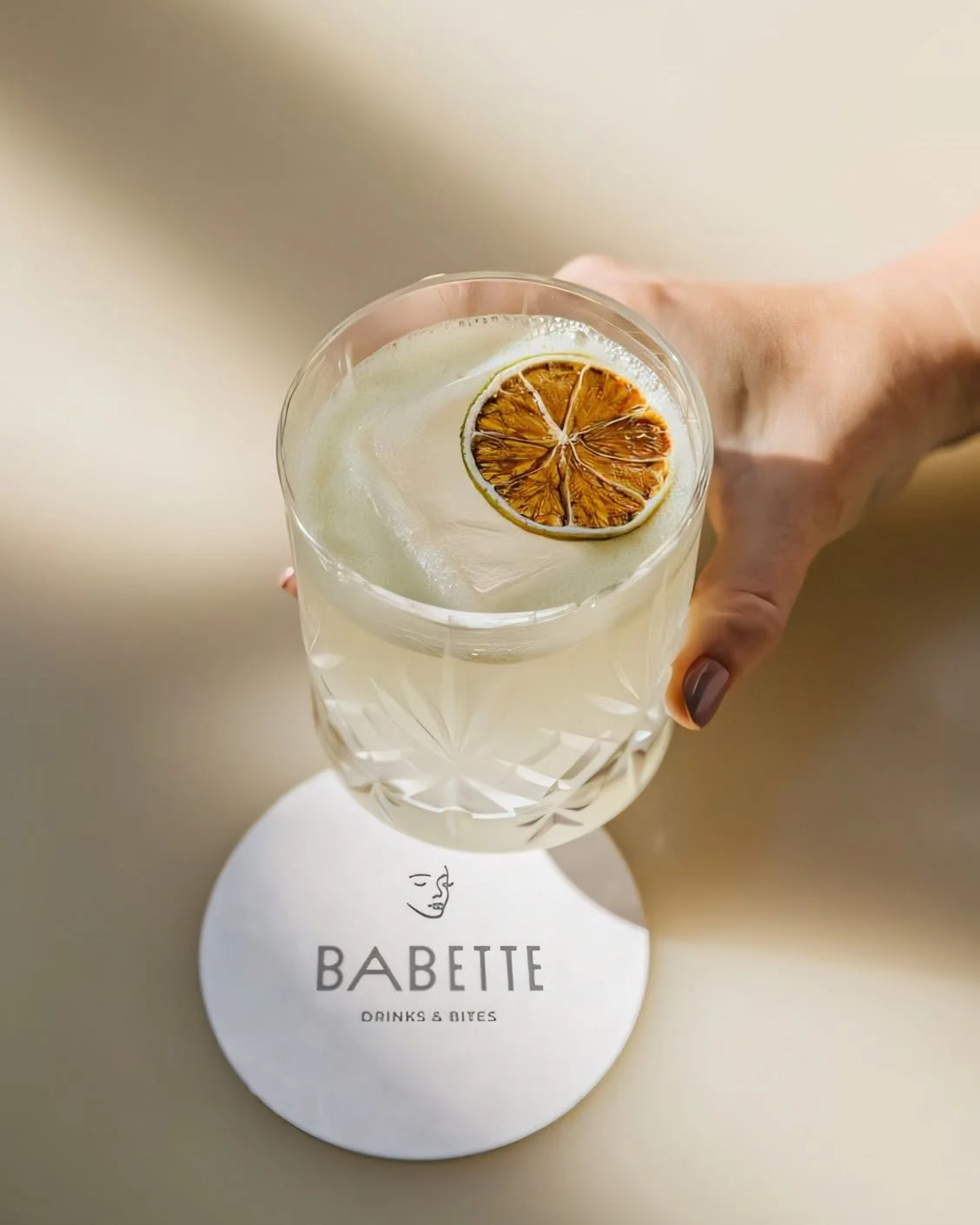 Een hand houdt een glas met een lichte cocktail, belegd met een gedroogd citroenschijfje, boven een witte onderlegger met het logo 'Babette' en de tekst 'Drinks & Bites'.
