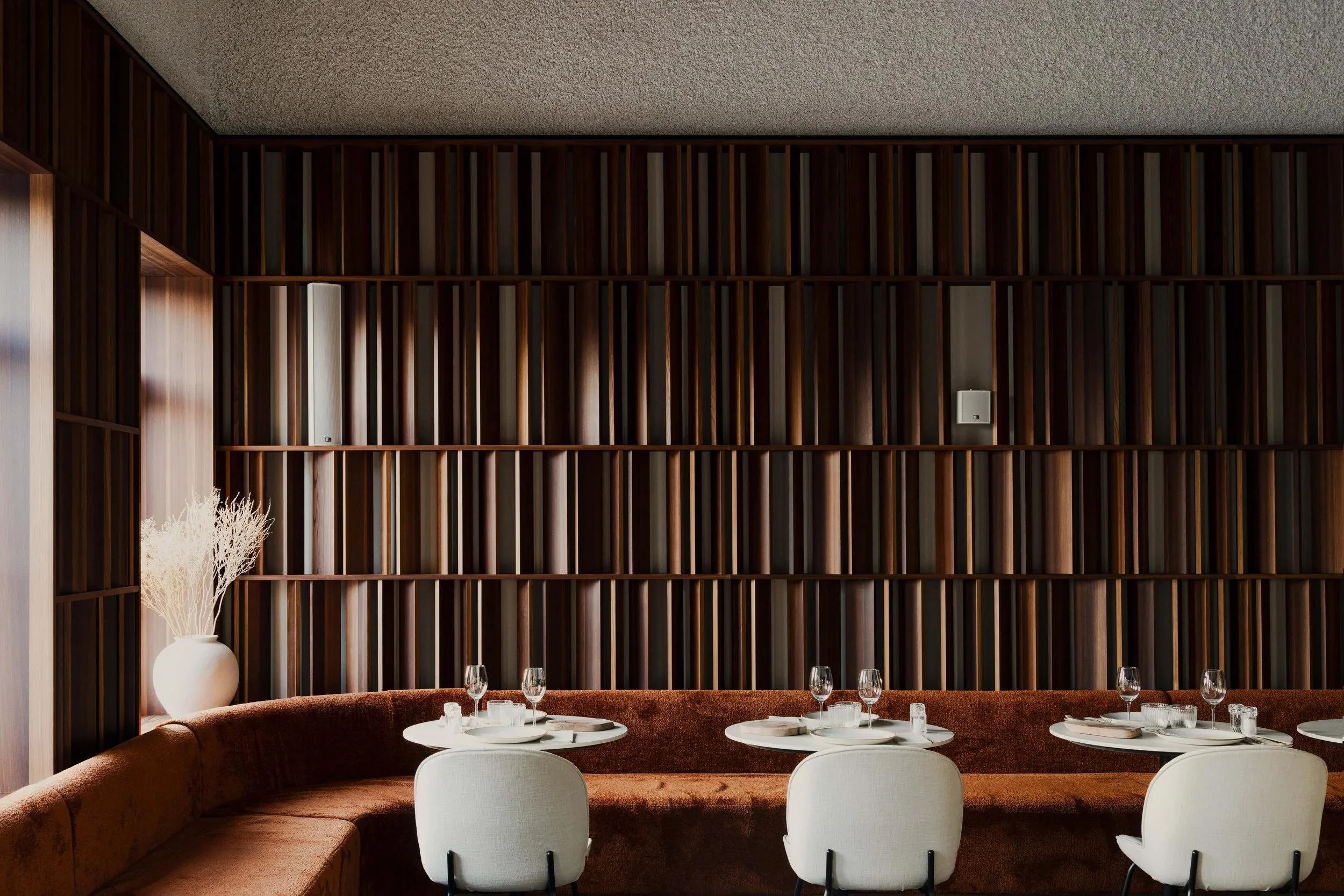 Een elegant restaurant interieur met een grote houten boekenplank, ronde witte tafels met wit servies en glazen, en witte stoelen.
