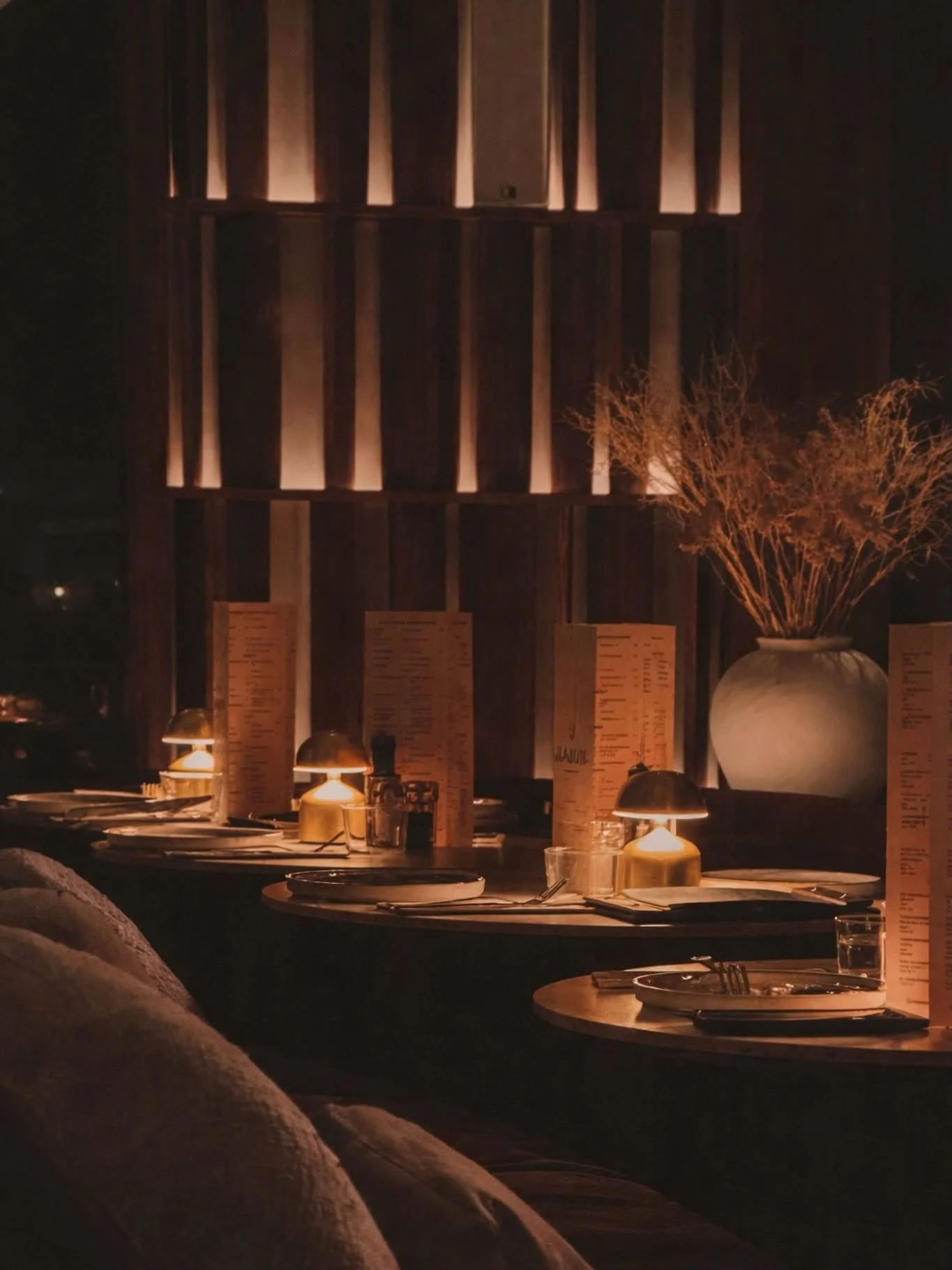 Een gezellige avond in een restaurant met gedimde verlichting, ronde tafels gedekt met borden, bestek en wijnglazen, en grote vazen met gedroogde takken op de achtergrond.