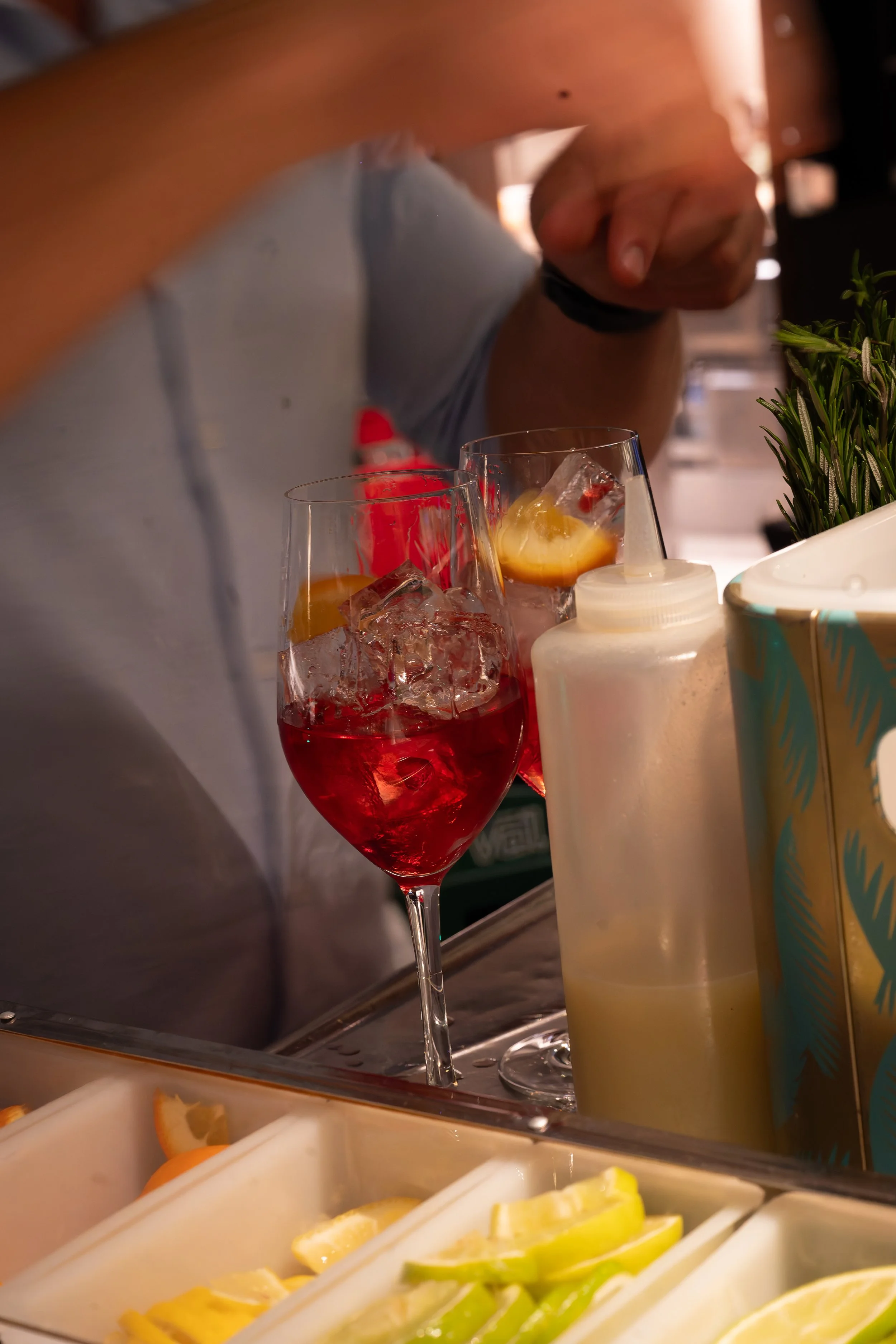 Een cocktailglas gevuld met een rode drank, ijsblokjes en een schijfje citrusfruit, op een bar of servingstation.