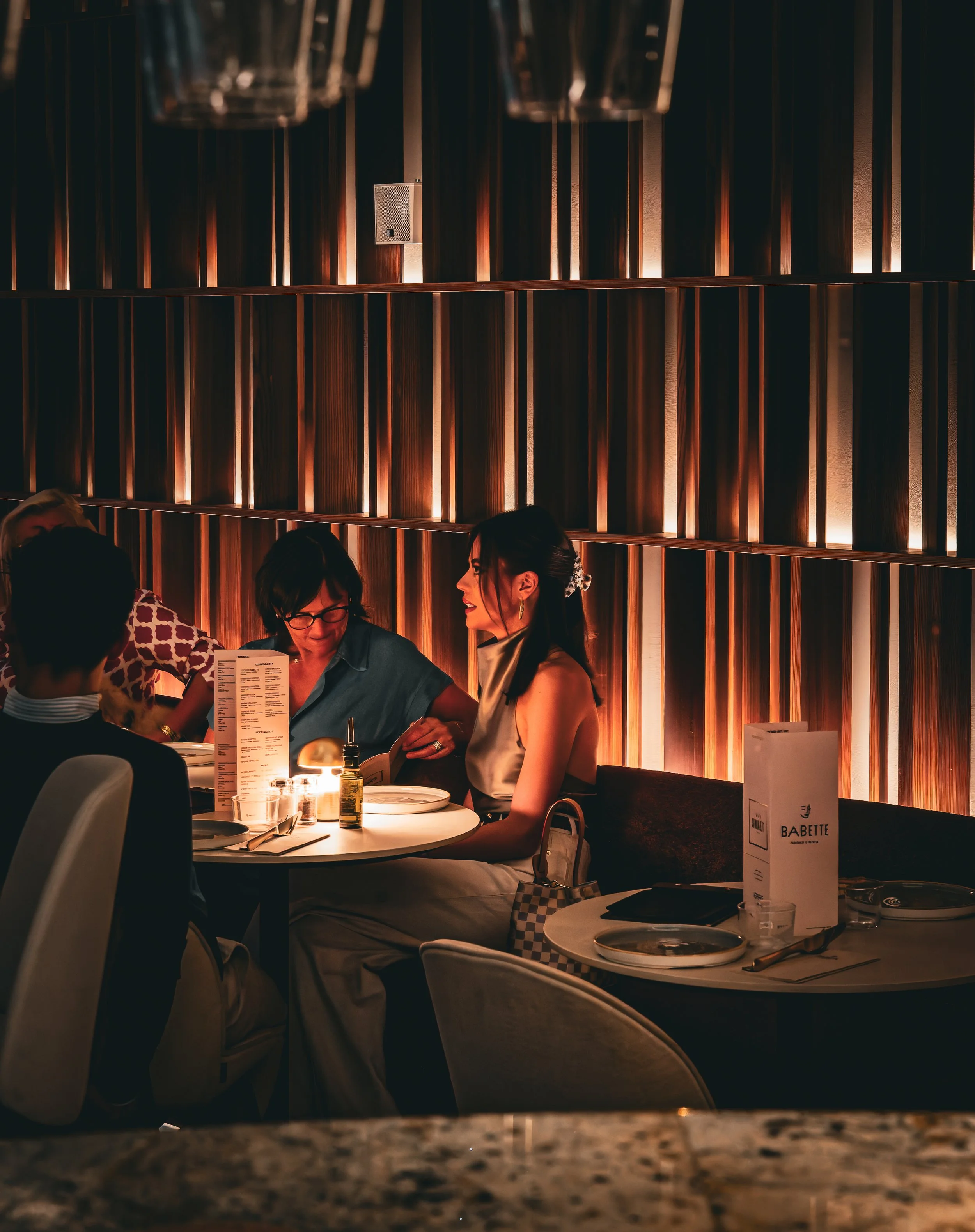 Mensen zitten aan een tafeltje in een restaurant met warme, sfeervolle verlichting en een houten muur op de achtergrond.
