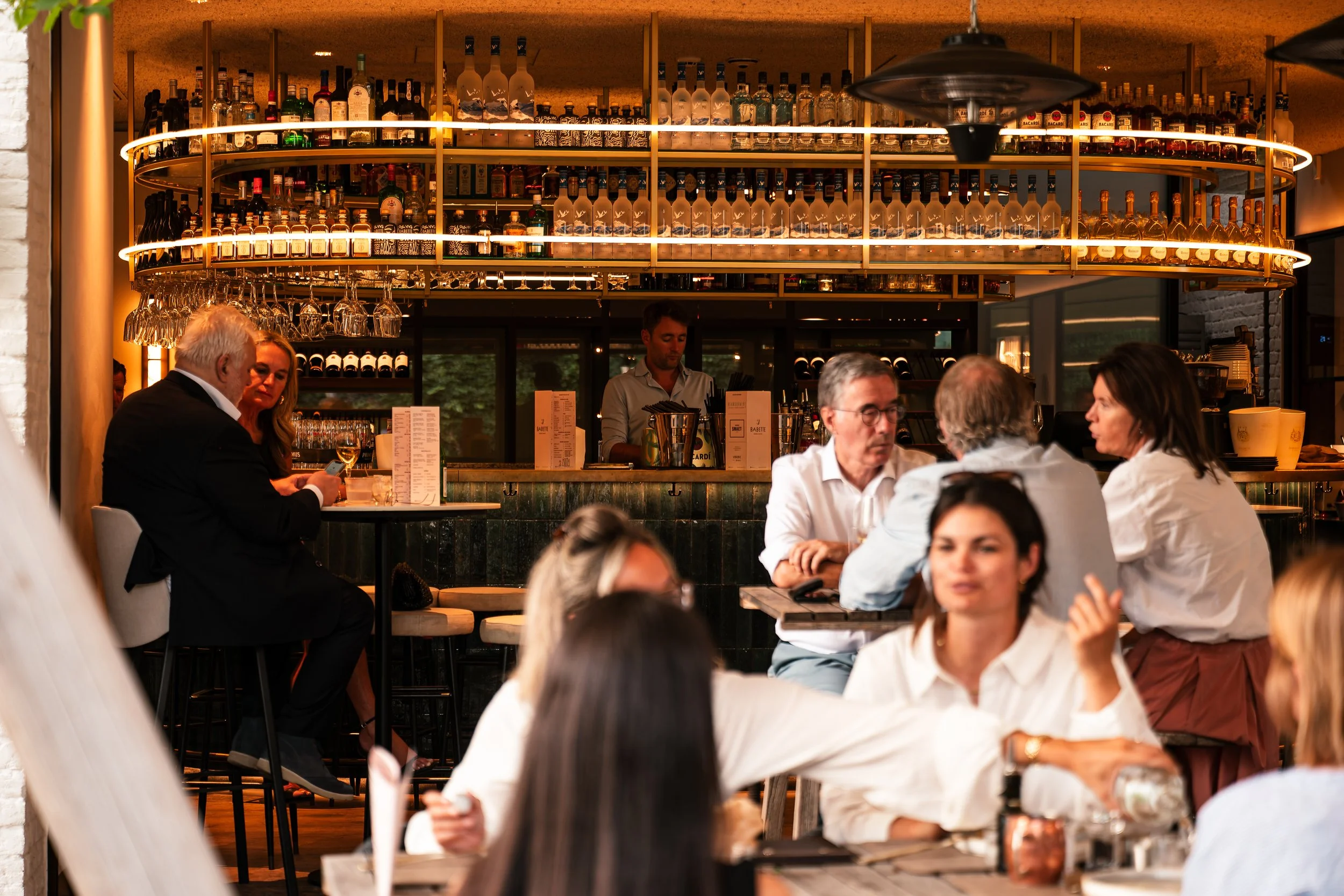 Binnen in een bar of restaurant met mensen die aan tafels zitten en met een bar waar verschillende soorten alcohol opgehangen en geserveerd worden.
