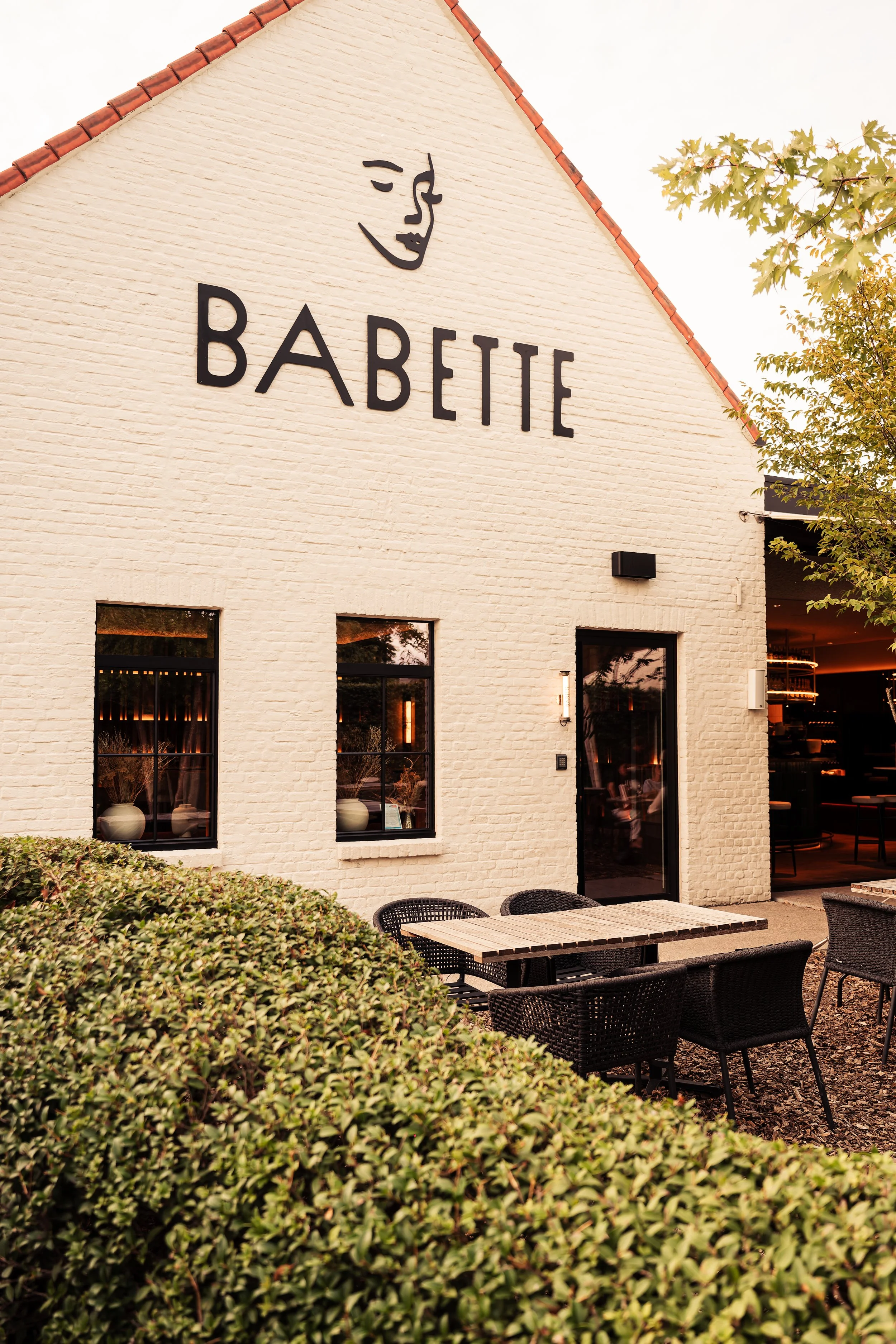 Een wit gekalkte gevel van een restaurant met de naam 'BABETTE' en een teken van een vrouw met gesloten ogen, omlijst door groene struiken en een terras met zwart wicker stoelen en een houten tafel.