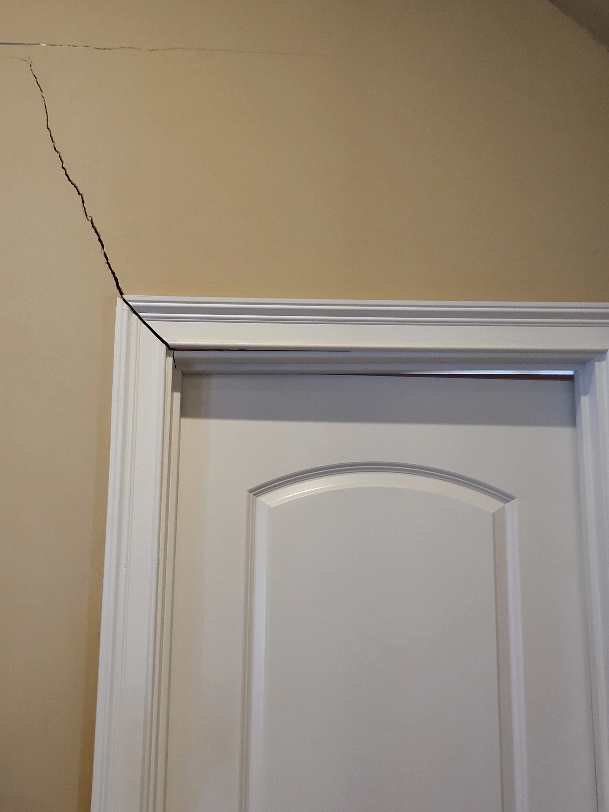 A white door with decorative panels and a white door frame, set in a beige wall. A large crack runs along the upper left corner of the wall.