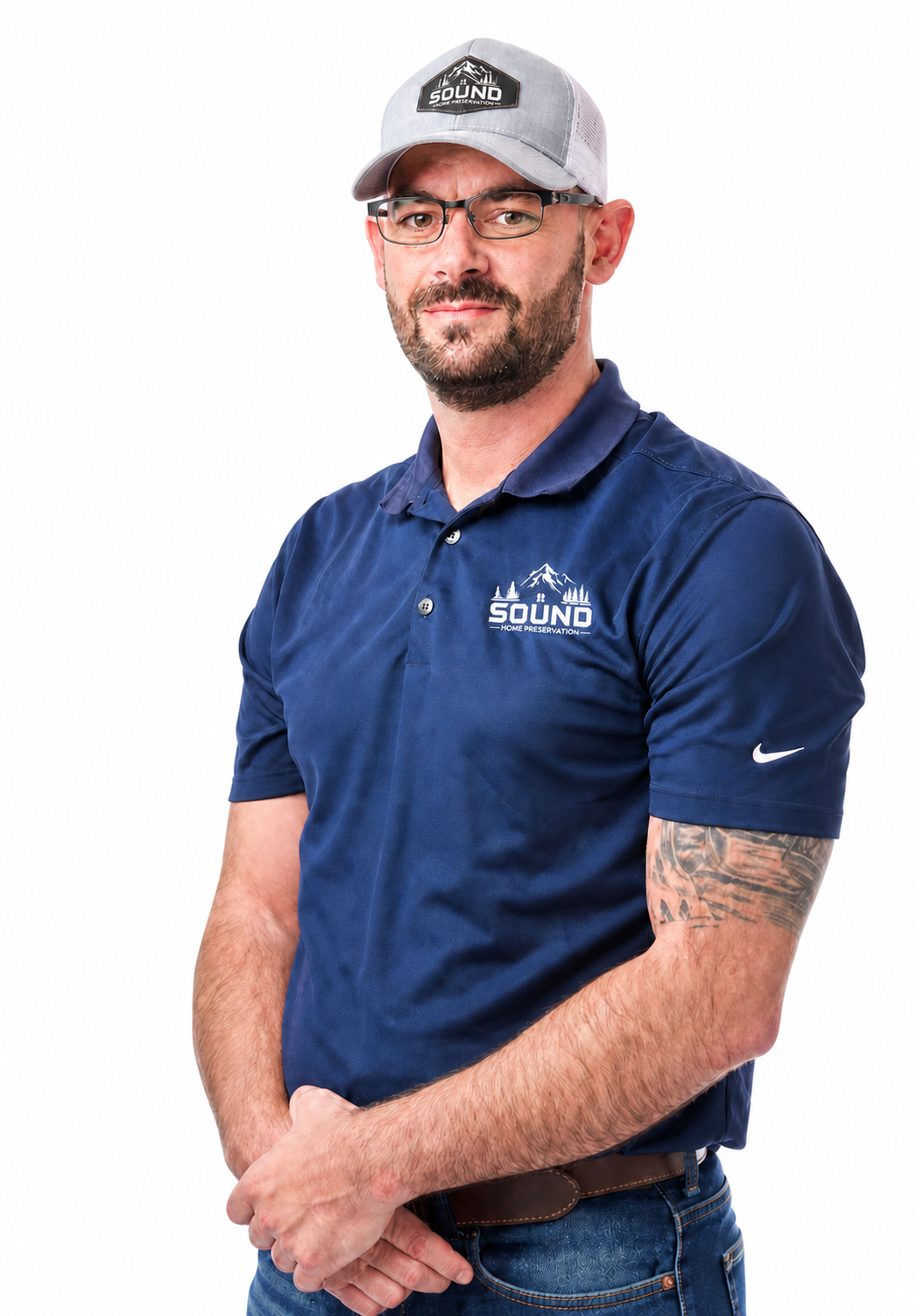 A man wearing glasses, a baseball cap, and a navy blue polo shirt with a logo that says 'Sound Home Preservation' on a mountain and trees background. He has a tattoo on his right arm and is standing against a plain white background.
