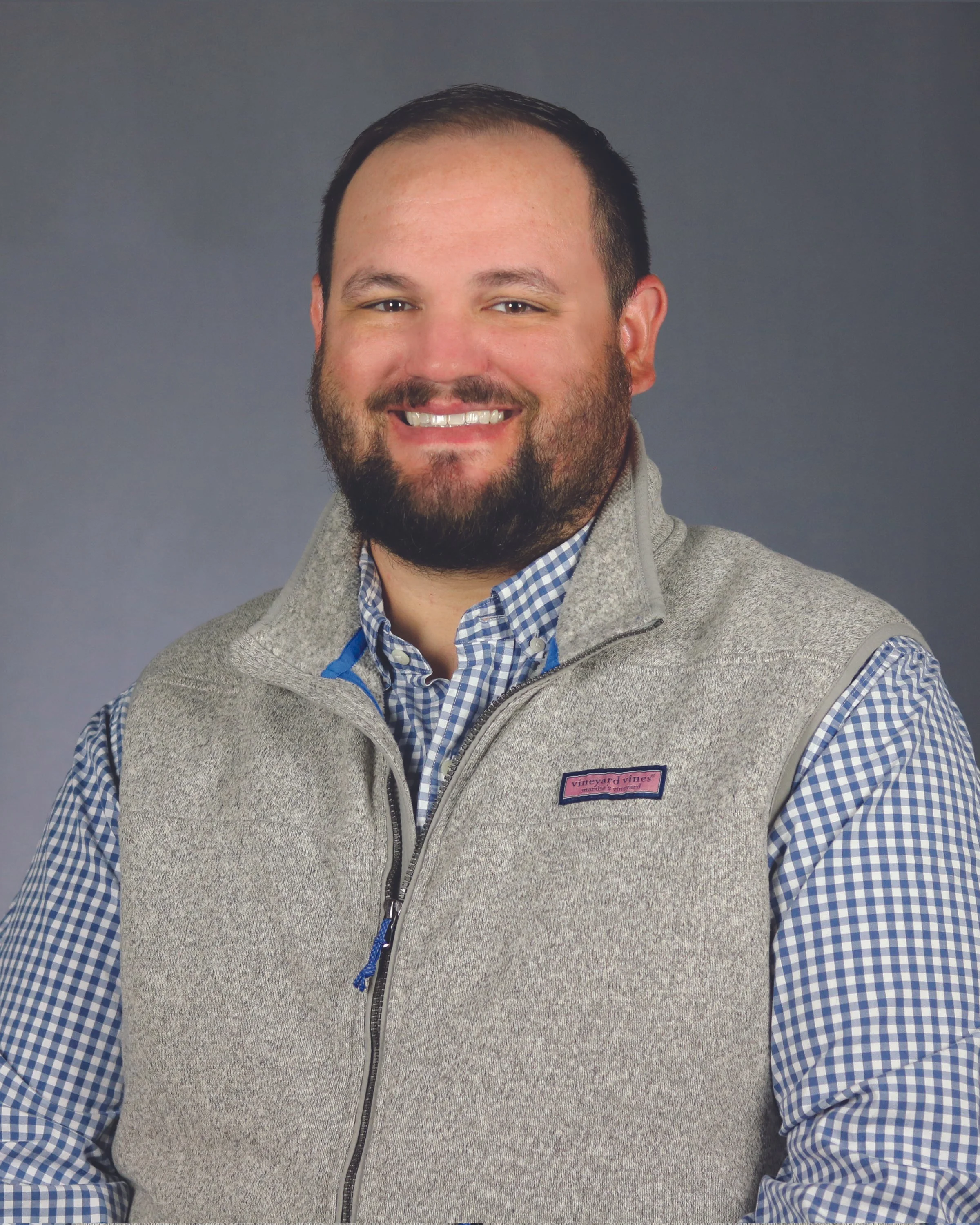 Close-up of a smiling man with a beard, wearing a gray vest over a blue and white checkered shirt, against a plain gray background.