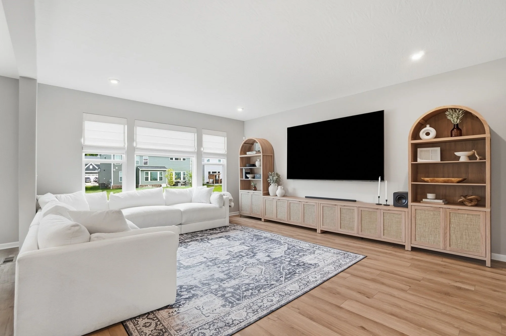 Living room with white L-shaped sofa, large windows, patterned rug, and built-in wooden shelves with decorative items, a mounted flat-screen TV, and hardwood flooring.