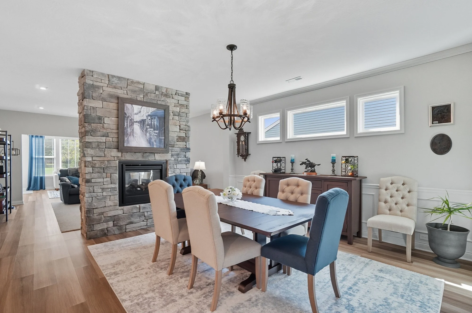 Open concept living and dining room with a stone fireplace, colorful chairs around a dining table, and large windows letting in natural light.