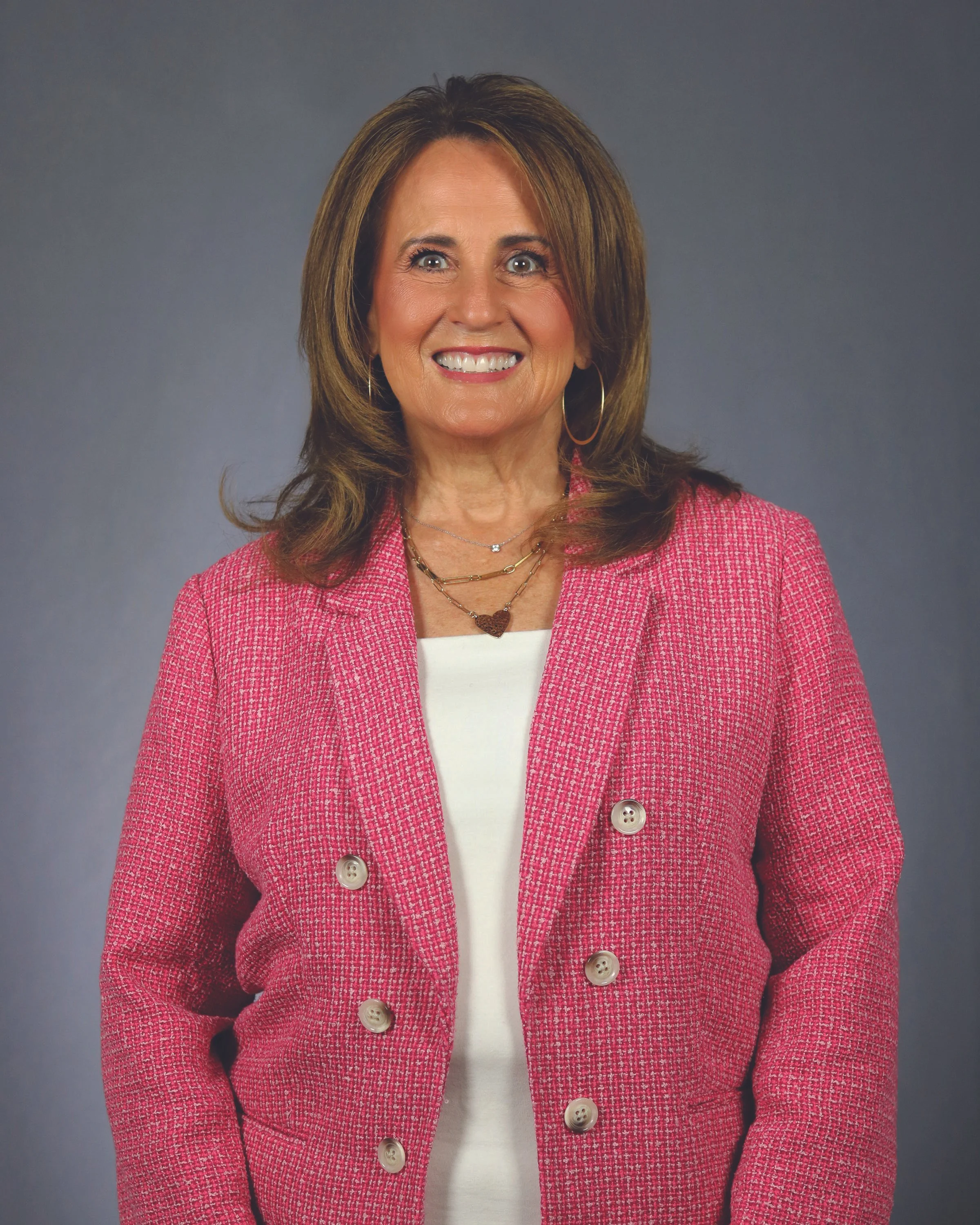 A woman wearing a pink textured blazer with large buttons, layered necklaces, hoop earrings, and a white top, smiling against a gray background.