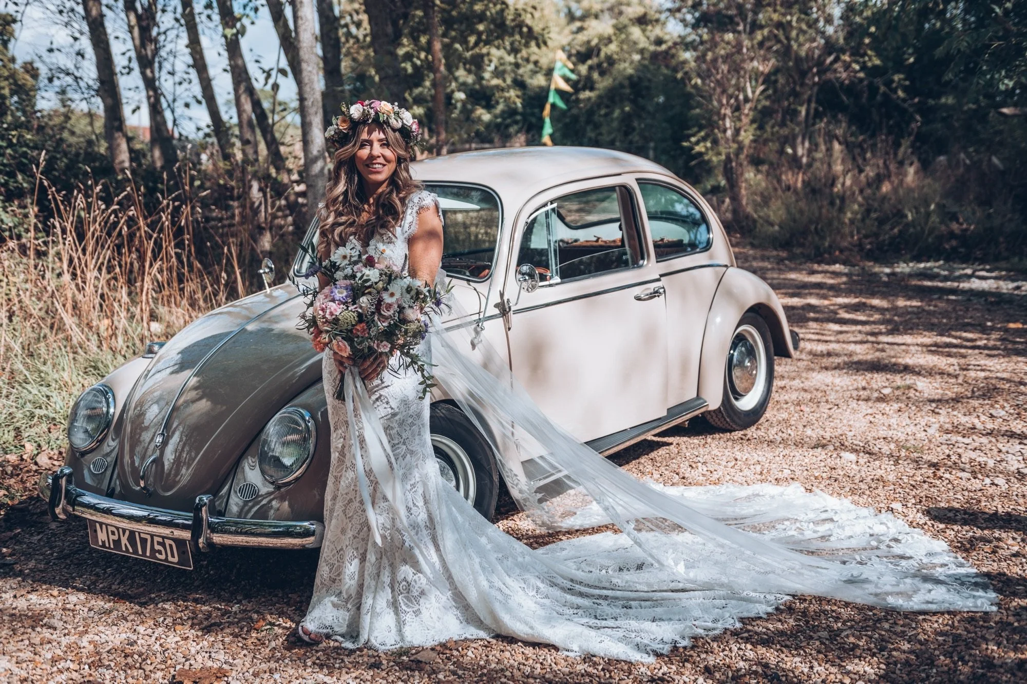 A smiling woman in a wedding dress with a flower crown and bouquet, standing next to a vintage Beige VW beetle car in a wooded outdoor setting.