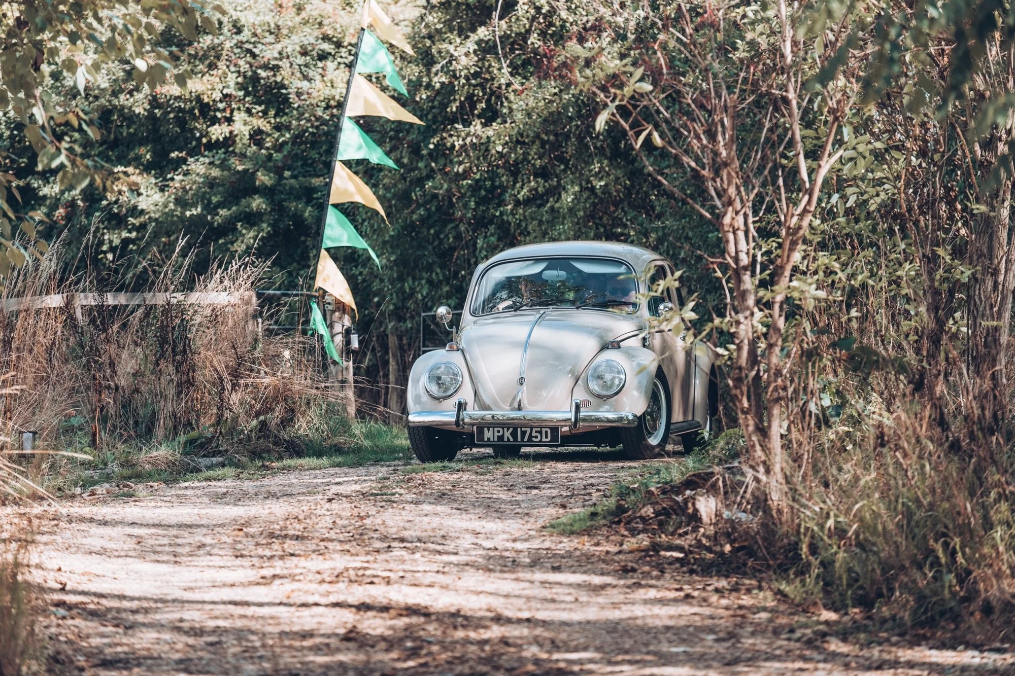 A vintage savanna Volkswagen Beetle parked on a dirt path in a wooded area, with trees and greenery surrounding it and some decorative flags hanging nearby.