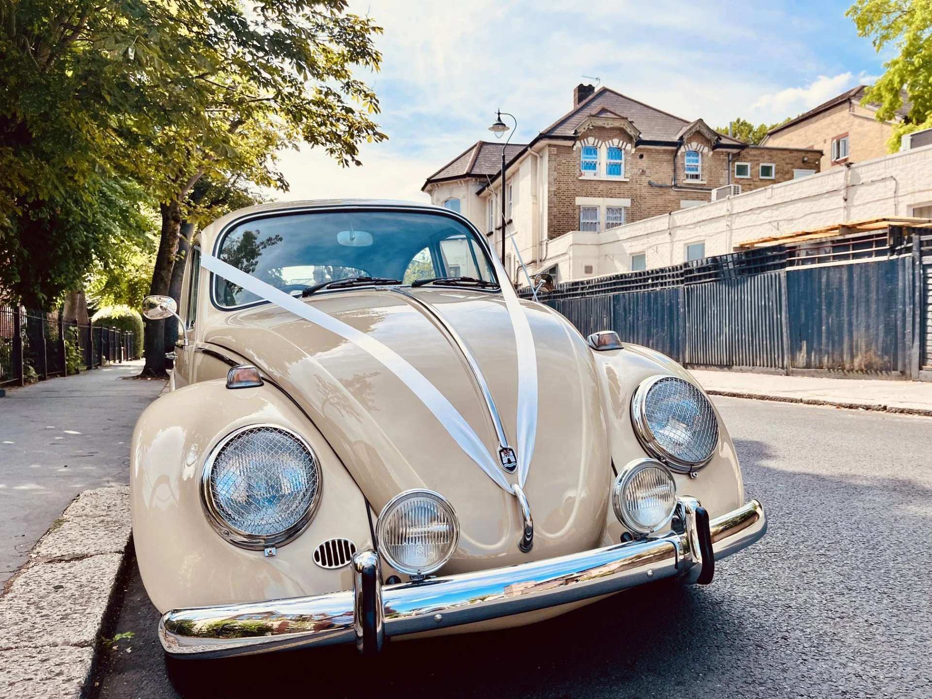 A vintage cream-colored Volkswagen Beetle parked on the street with trees, a black metal fence, and residential buildings in the background.