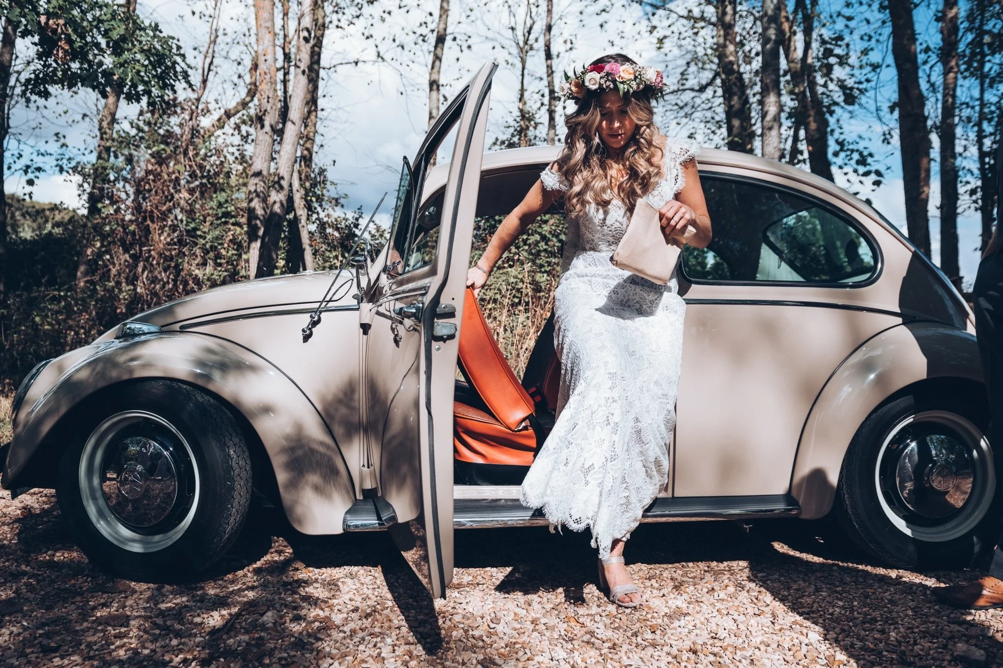 A beautiful bride wearing a white lace wedding dress and a floral headpiece steps out of a vintage beige car in a forest setting.