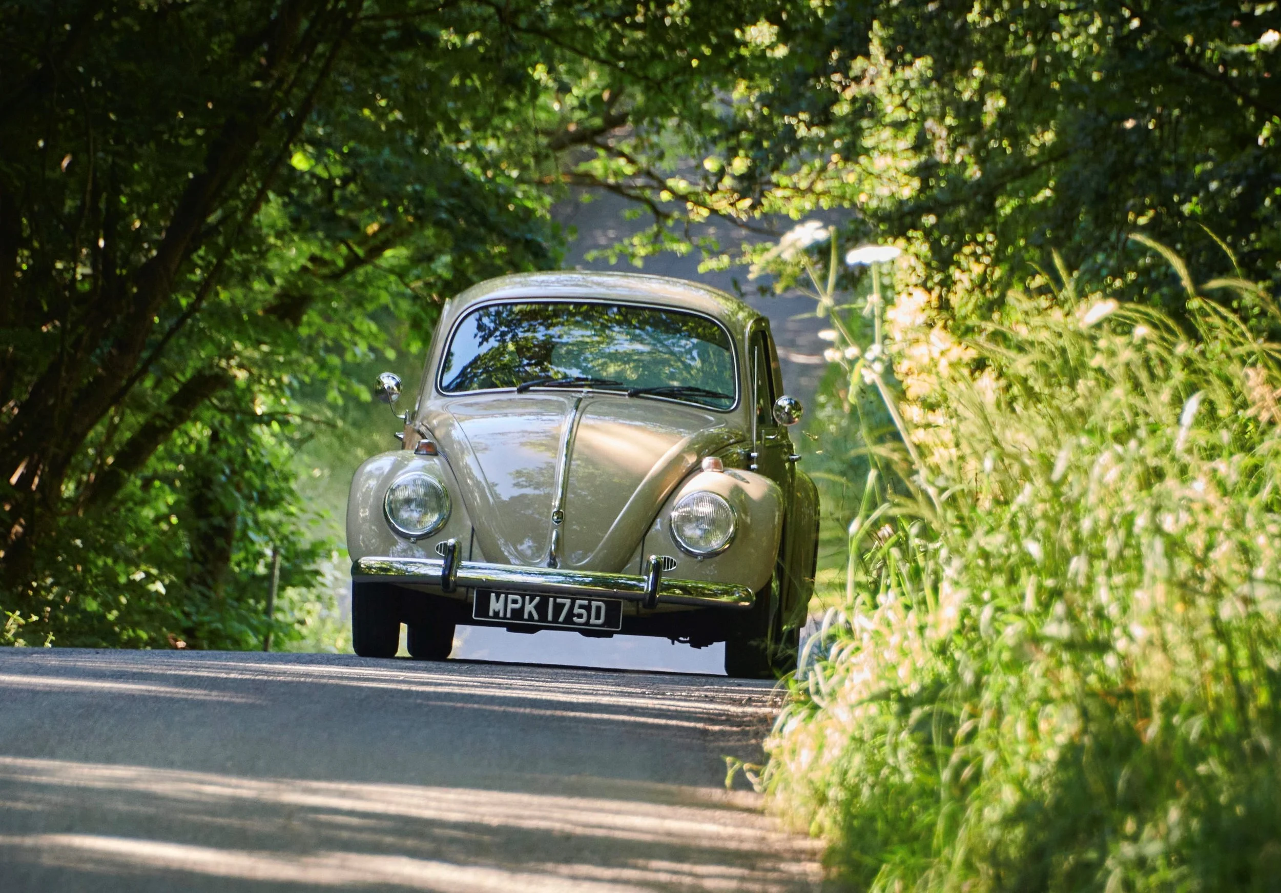 A vintage beige VW beetle car driving down a narrow country road surrounded by lush green trees and foliage.