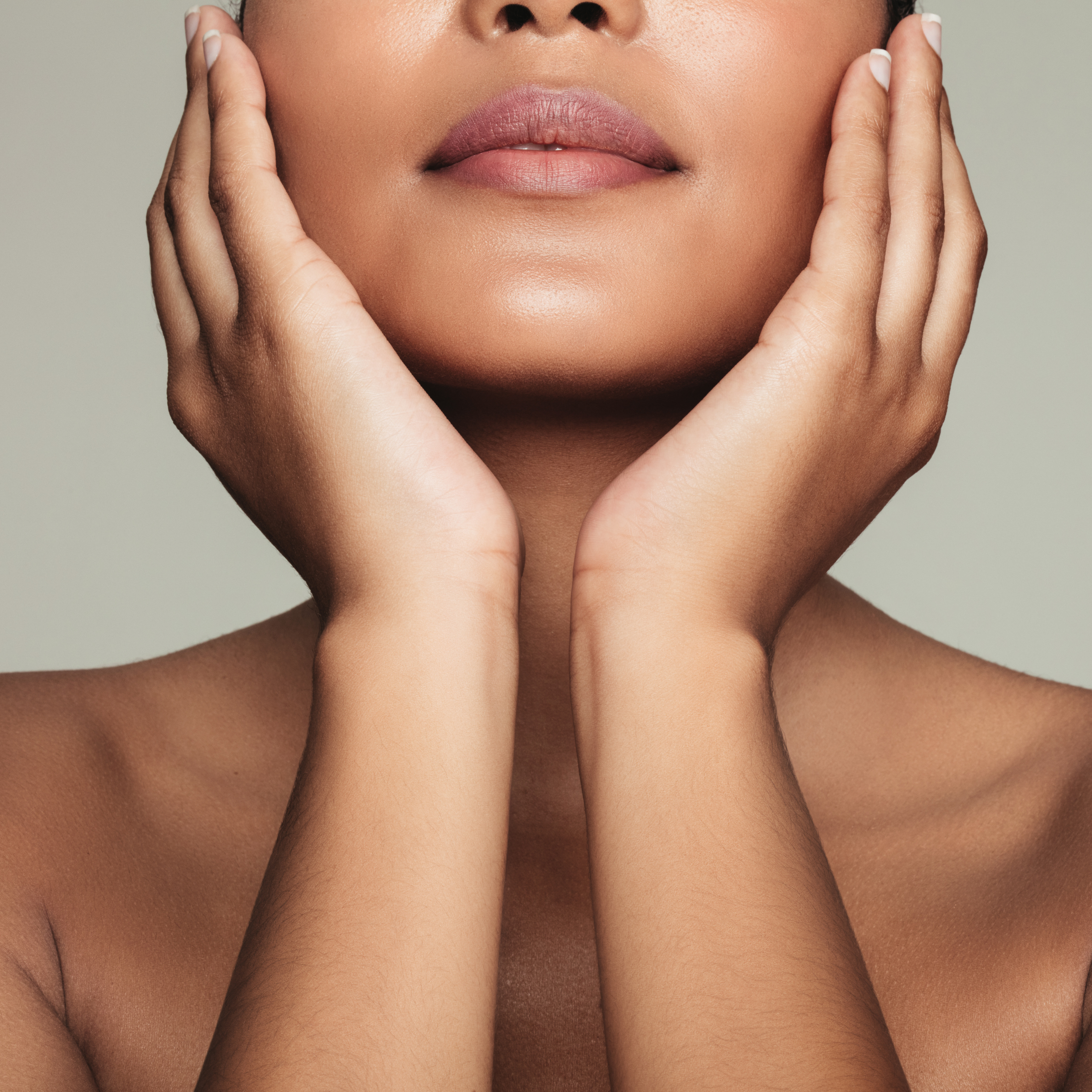 Close-up of a woman's face with hands framing her cheeks. She has smooth skin, full lips, and slightly parted mouth. The background is plain and neutral-colored.