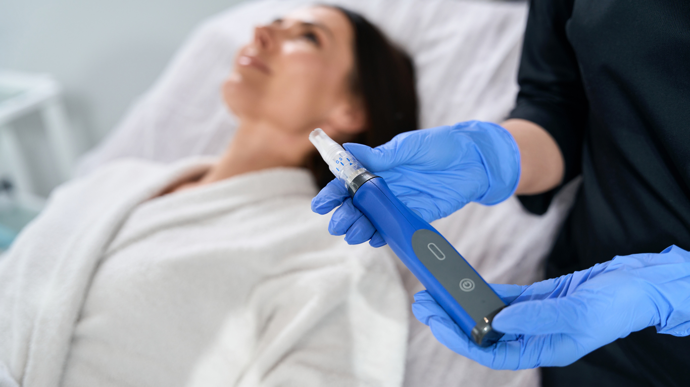 An aesthetician preparing to perform microneedling to a woman lying in a hospital bed, holding a microneedling device
