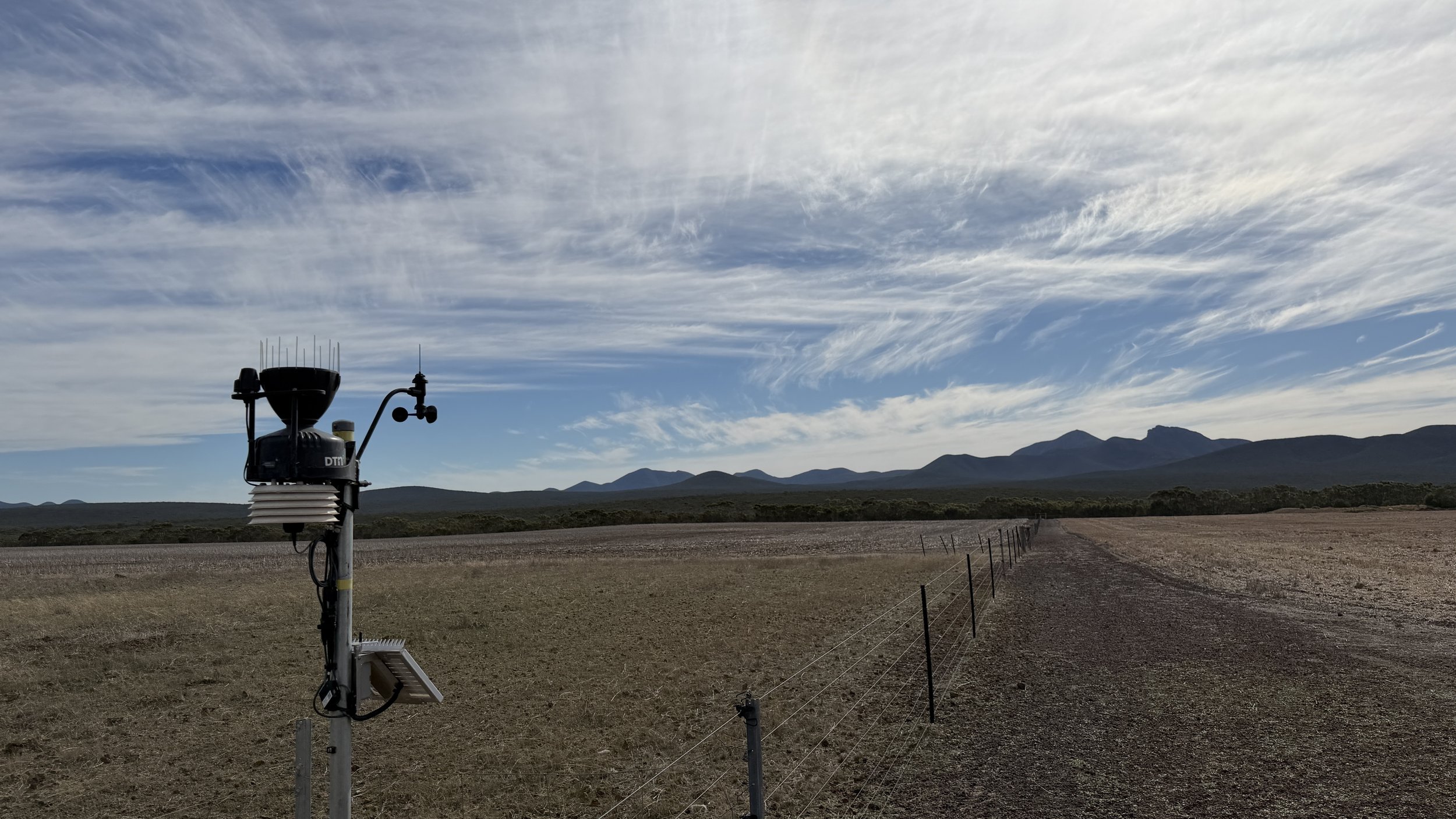 Weather station in a rural field with mountains in the background and a partly cloudy sky.