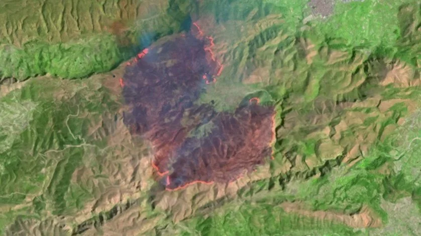 Satellite image of a large forested area with a wildfire evident by dark smoke and burnt land along the fire perimeter in the center.