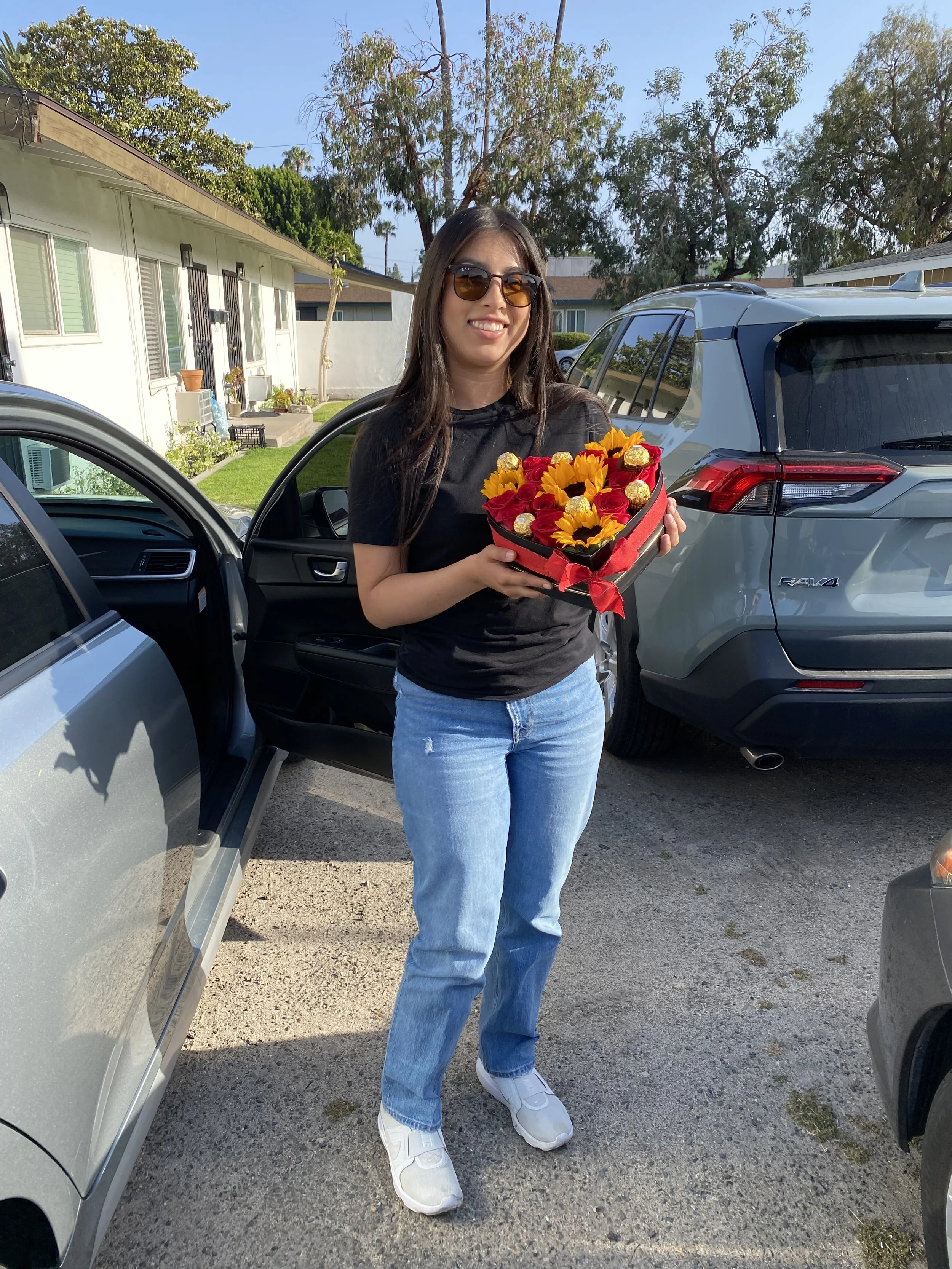 A woman with long dark hair wearing sunglasses, a black t-shirt, and light blue jeans, smiling and holding a gift box of sunflowers and red roses in a residential driveway.