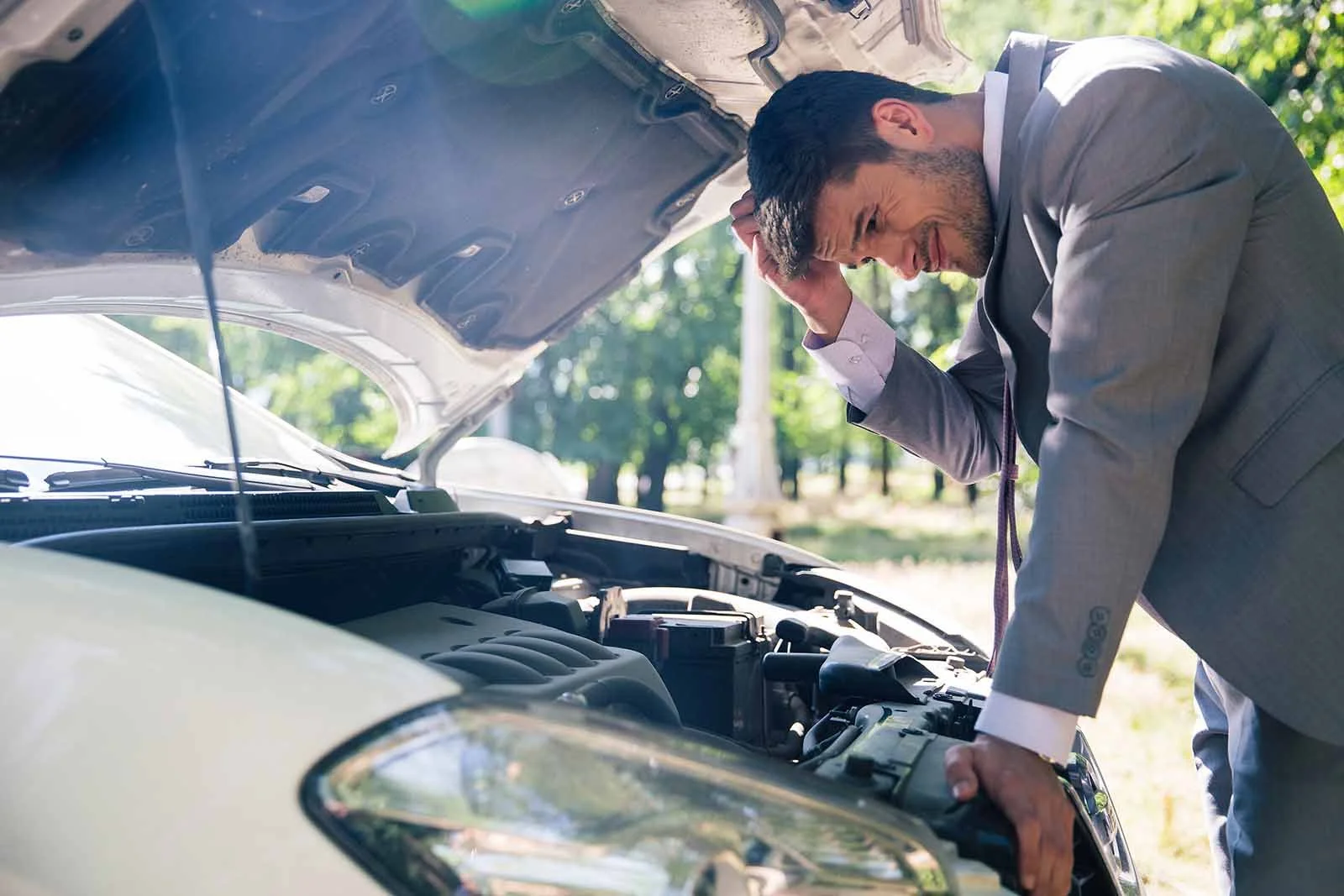 A man in a gray suit with a pink dress shirt and tie, looking distressed as he leans over the open hood of a car, inspecting the engine.