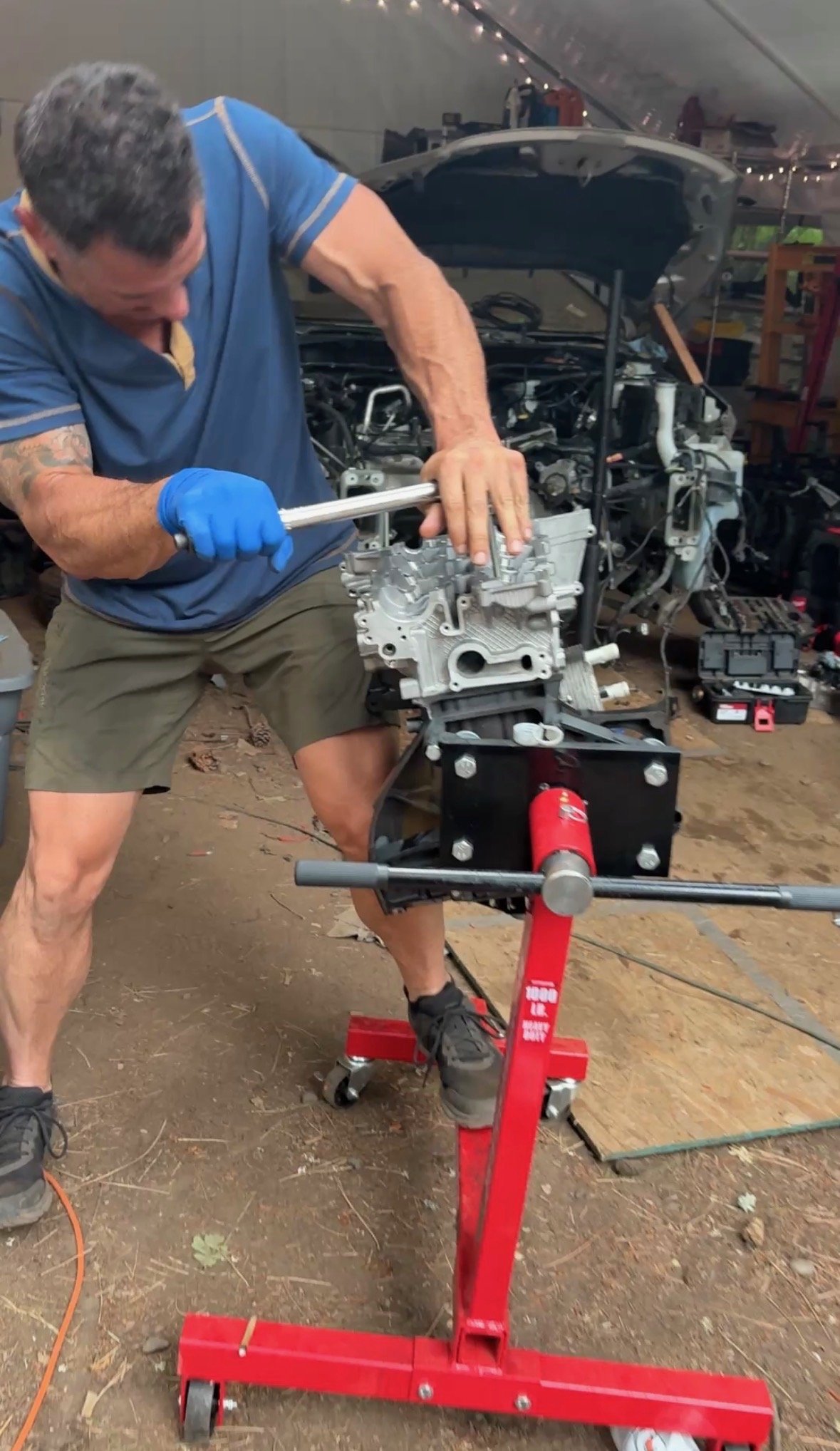 A man wearing a blue shirt, shorts, and gloves working on an engine mounted on a red engine stand inside a garage or workshop.