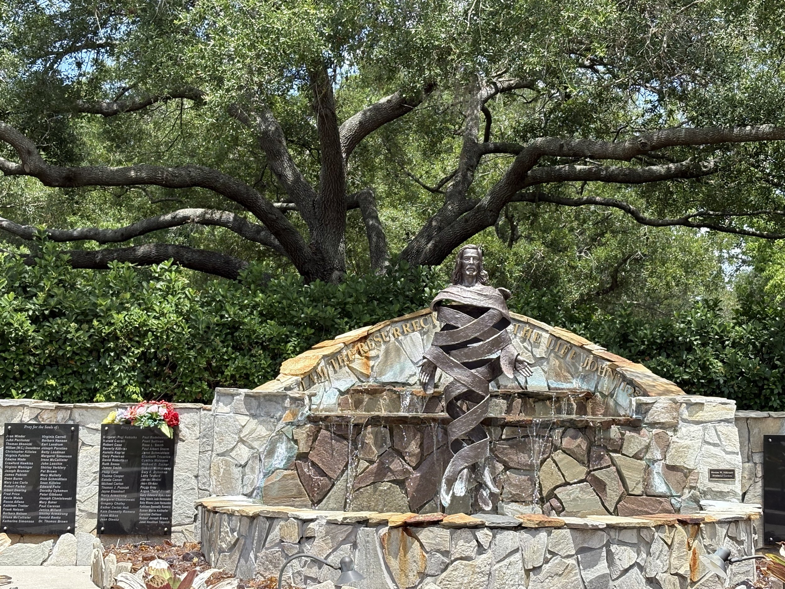 Memorial with a statue of Jesus Christ in front of a large leafy tree. The memorial features a stone wall with engraved names and a plaque, surrounded by flowers and decorative rocks.