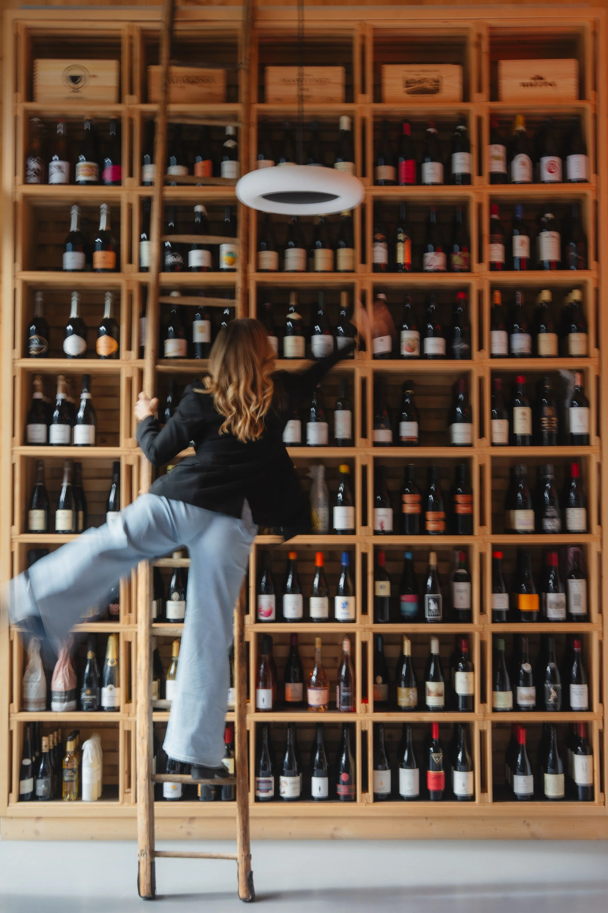 A person with long hair climbing a wooden ladder towards a wall of wine bottles arranged in wooden cubbies. The person is reaching for a bottle on the upper shelves.
