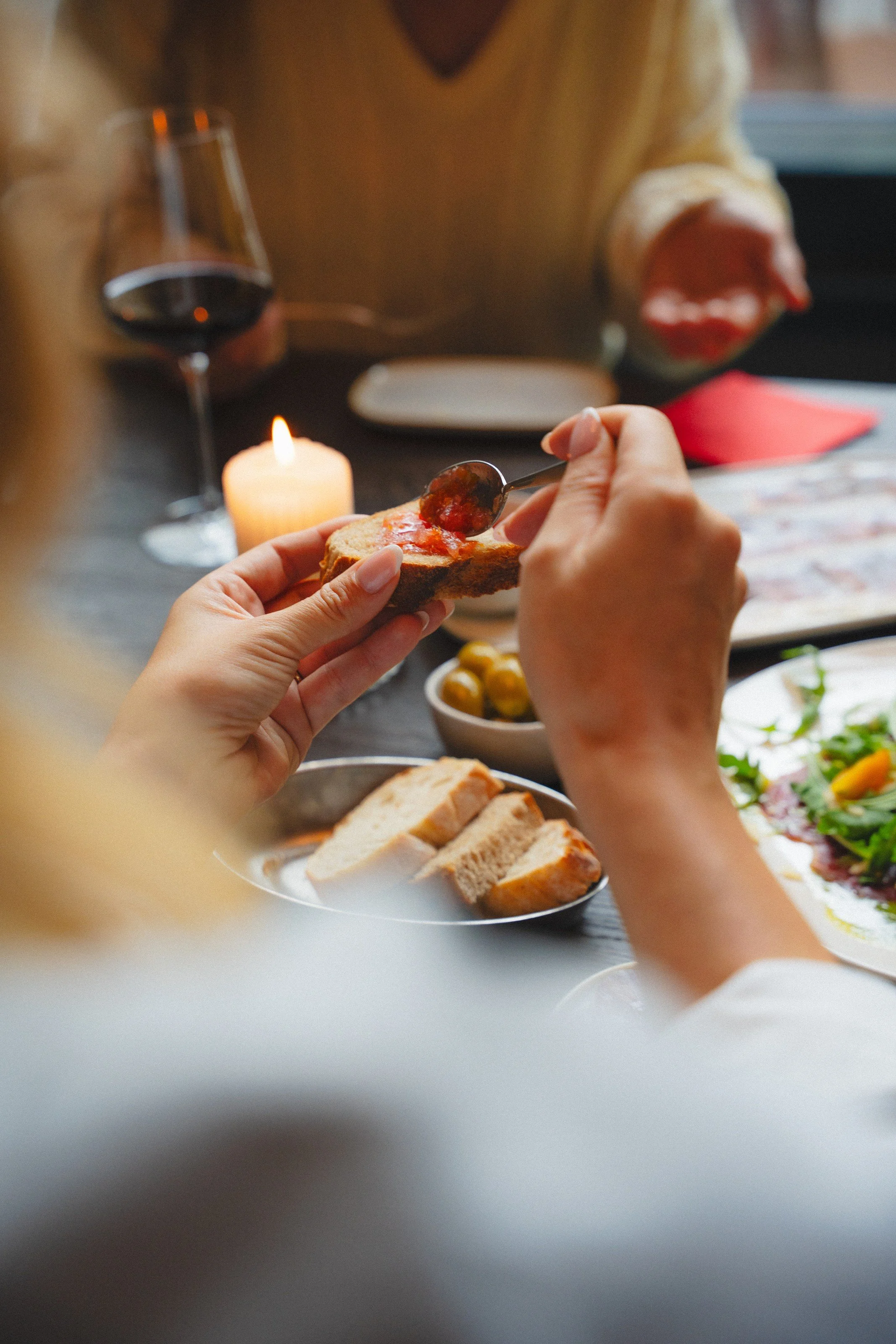 Person spreading jam on a slice of bread at a dinner table with a glass of red wine, candle, and assorted food dishes.