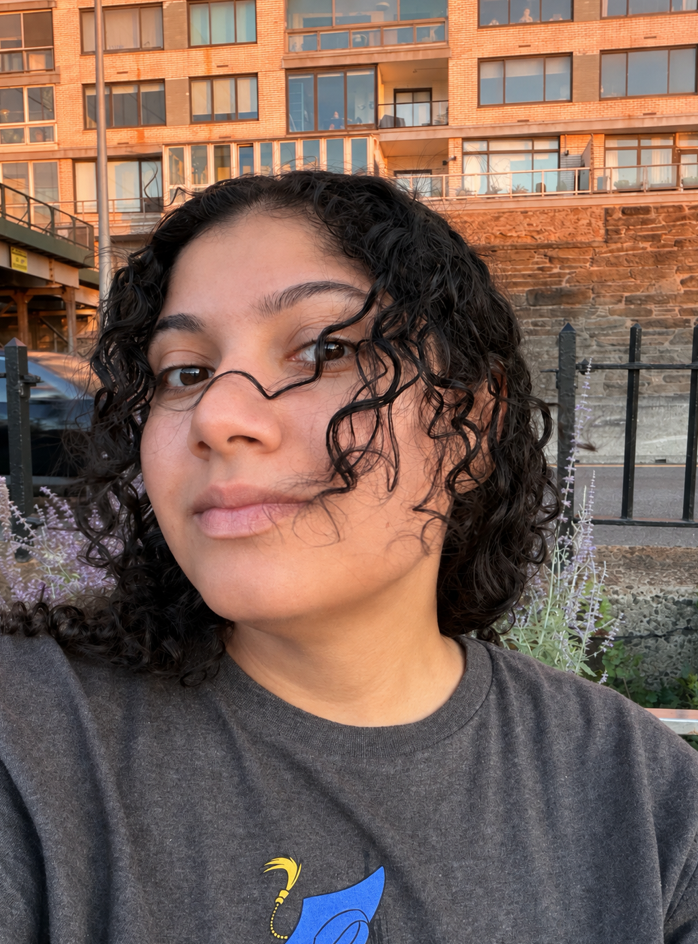 Close-up of a woman with curly dark hair, wearing a dark gray T-shirt, standing outdoors with an urban background featuring a brick and glass apartment building, a black metal fence, and purple flowers.