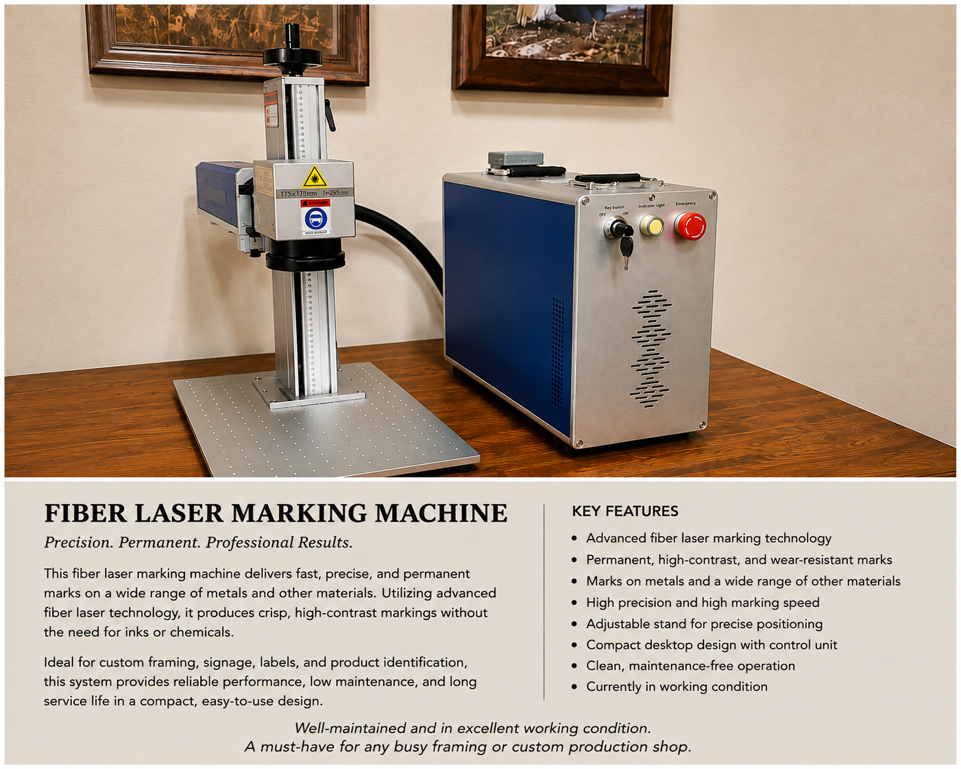 A fiber laser marking machine on a wooden table, featuring a tall vertical laser device connected to a blue and silver control unit with buttons for key switch, indicator light, and emergency stop. The background includes two framed paintings on a beige wall.