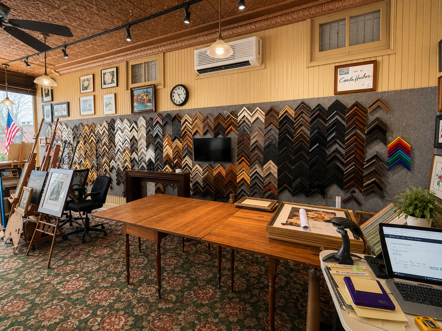 A store interior with a wall displaying various picture frame samples in different colors and sizes, a television, a clock, and framed artwork. There are tables, chairs, and art supplies, with a window showing an American flag outside.