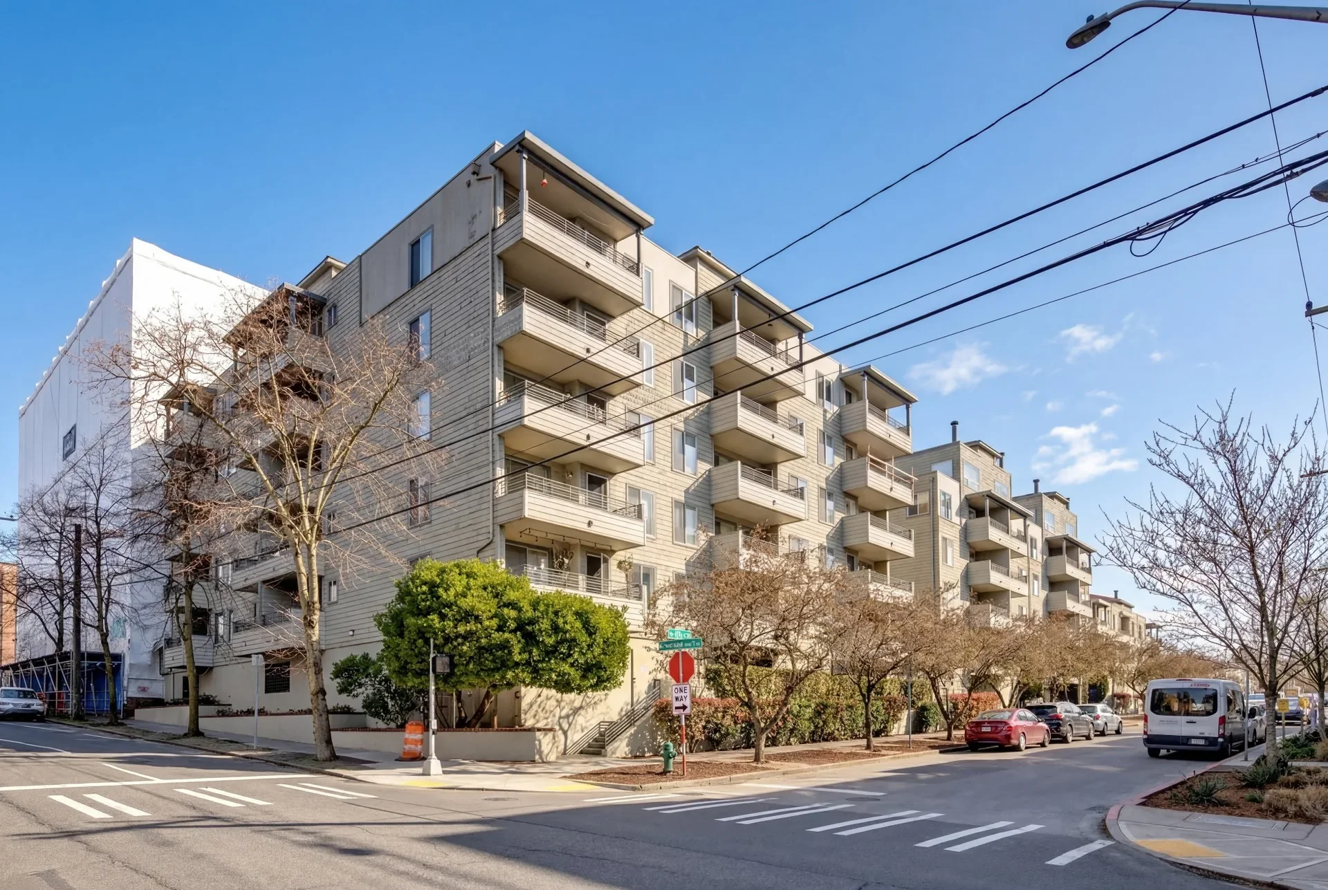 A multi-story apartment building with balconies and beige siding, situated along a street with trees and parked cars, under a clear blue sky.