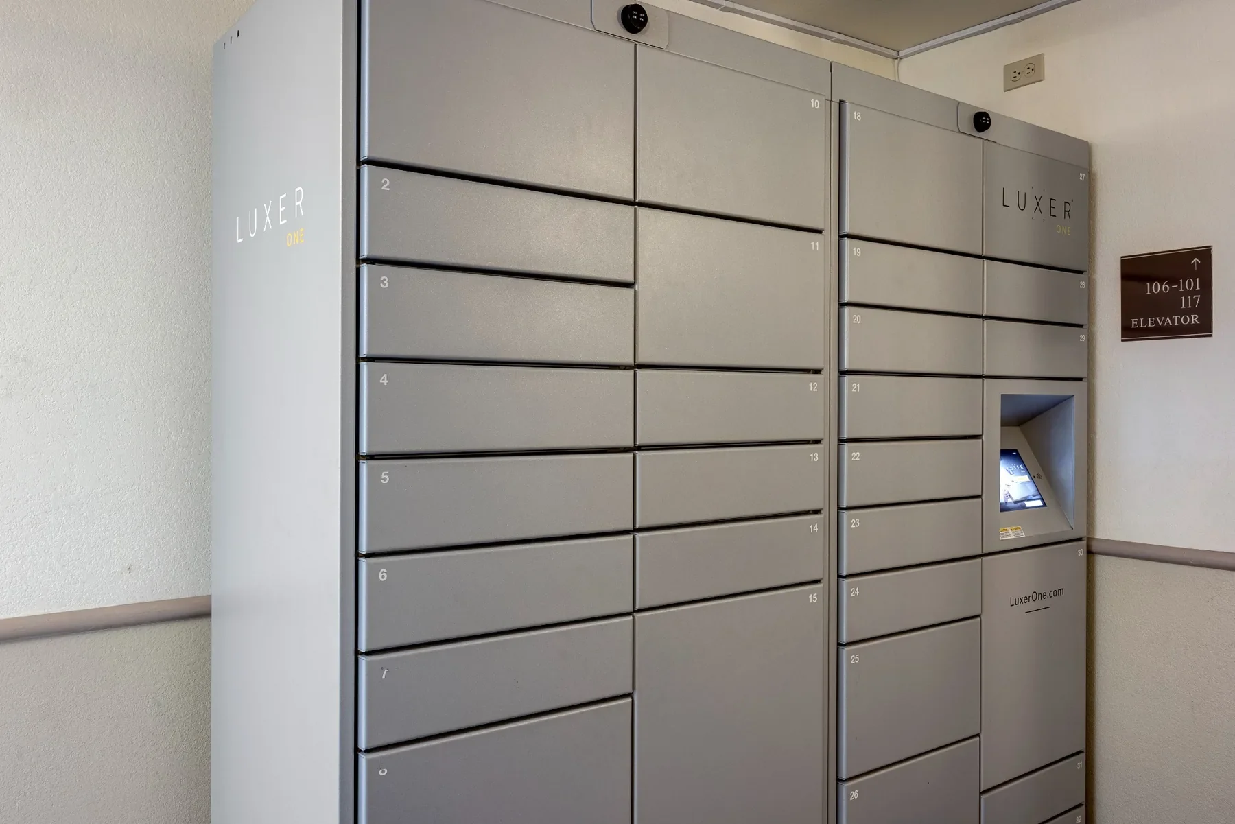 Gray parcel locker system labeled "Luxer One" with numbered compartments and an integrated touchscreen kiosk, located in a hallway near an electrical outlet and a sign indicating directions to the elevator.